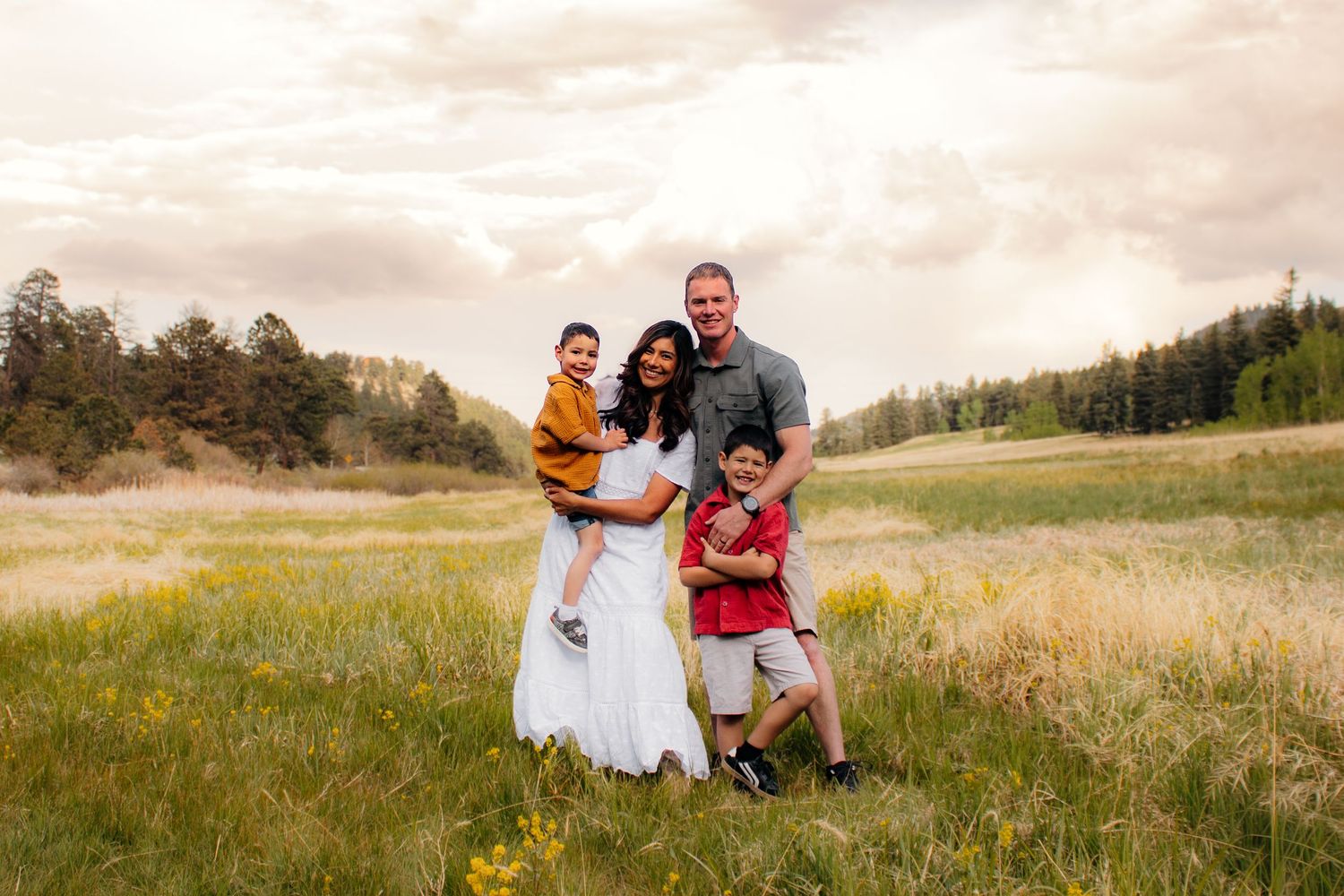 Happy family of four posing in a lush green meadow with wildflowers and pine trees under a dramatic cloudy sky.
