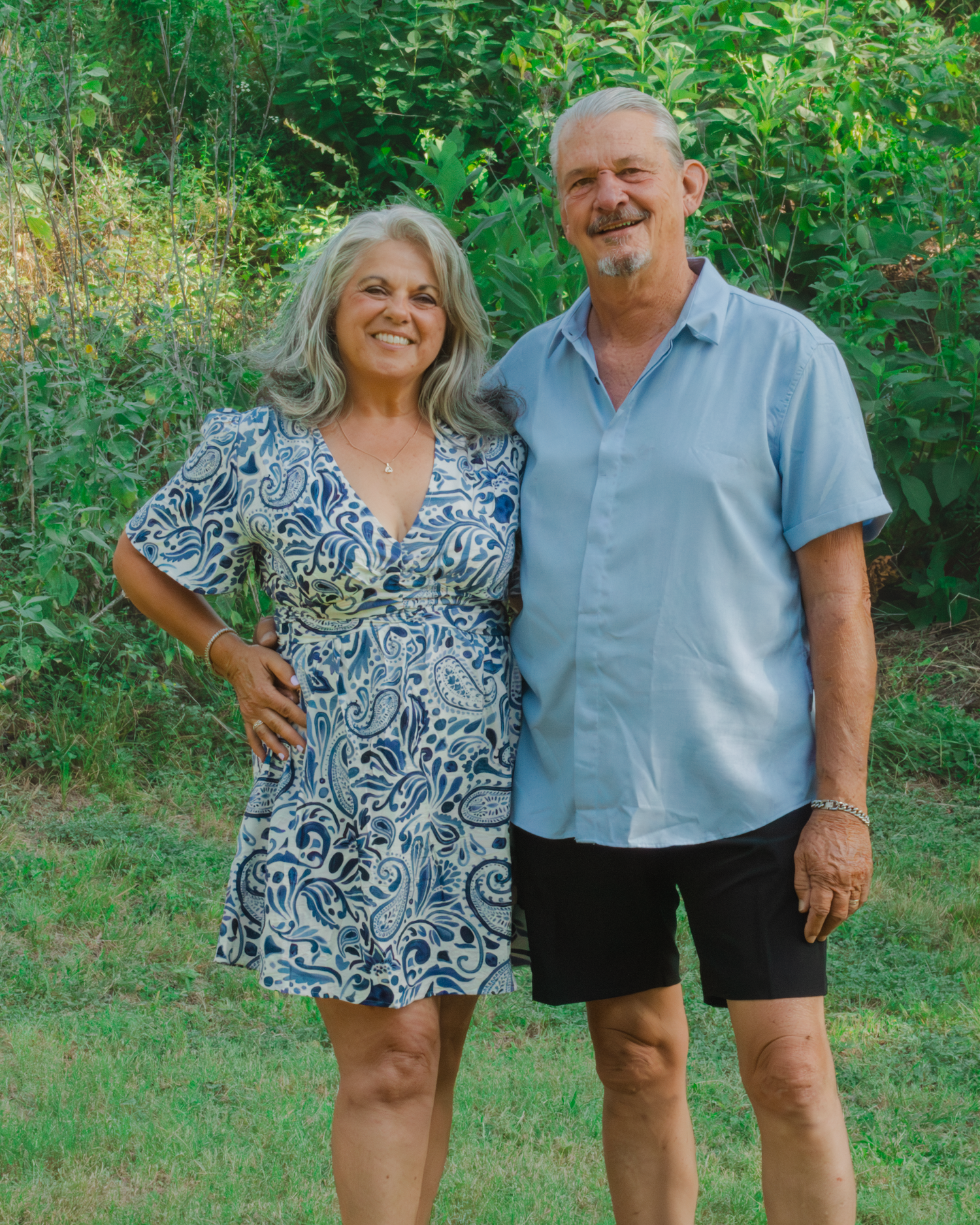 A smiling couple poses outdoors in a lush green garden, woman in blue paisley dress, man in light blue shirt and black shorts.