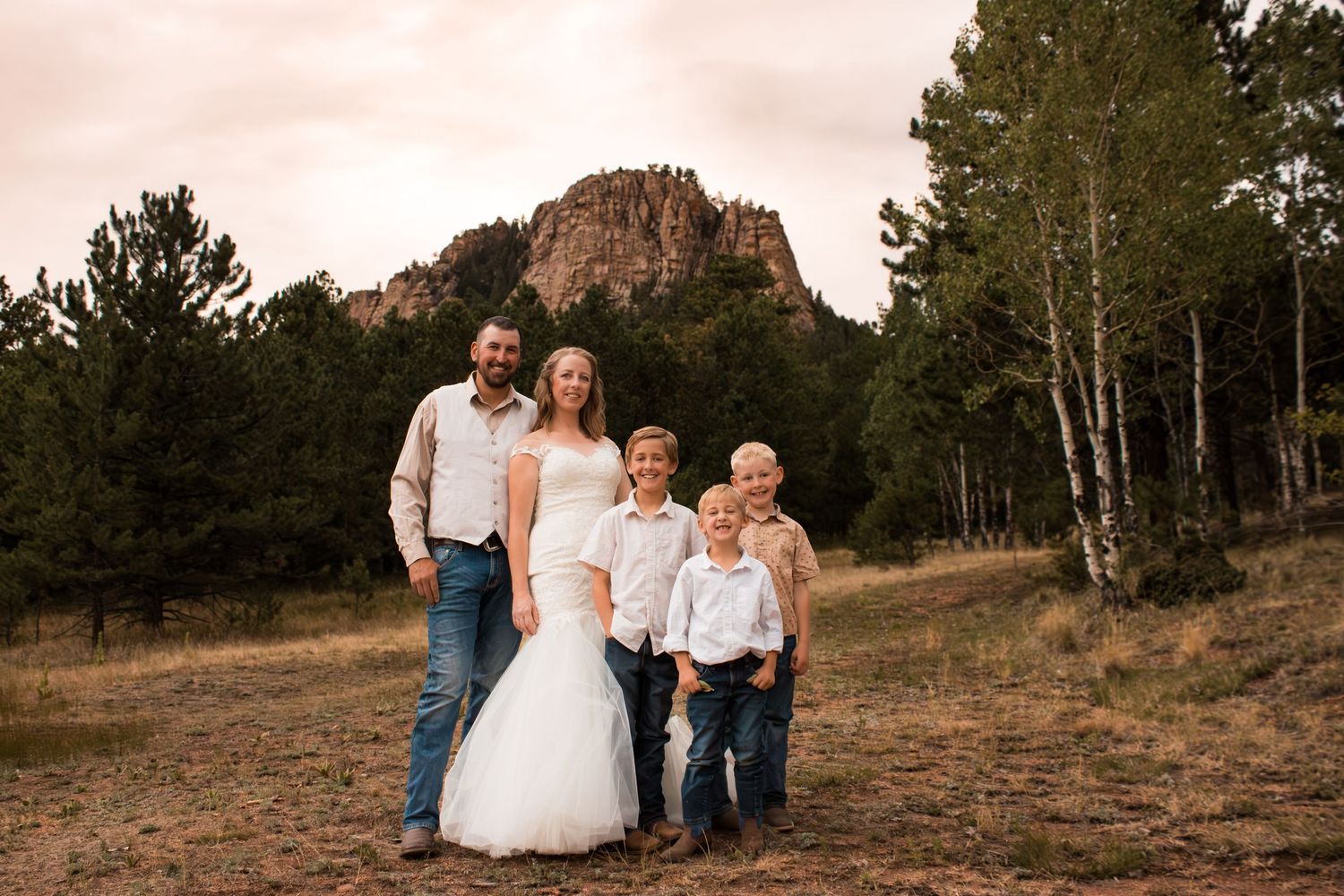 Family of five poses outdoors in wedding attire near pine trees and rocky mountain backdrop.
