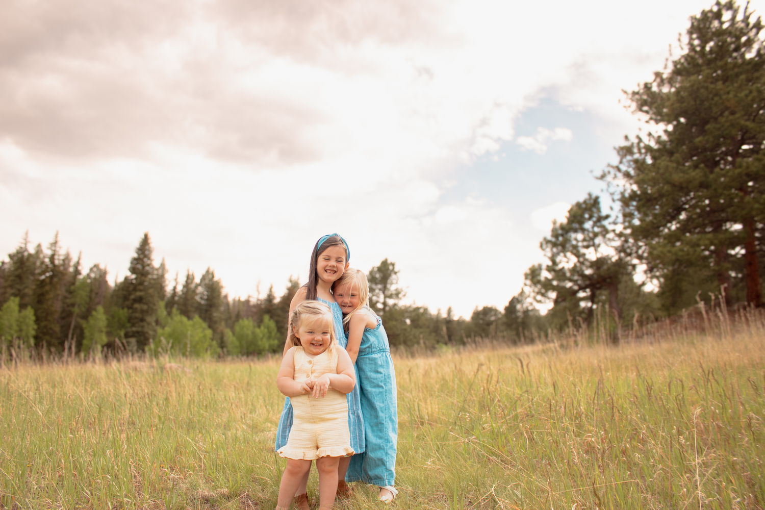 Three young sisters smiling together in a lush green meadow with tall grass and pine trees.