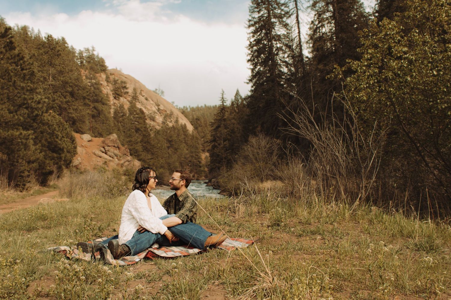 Couple sitting together on a blanket in a scenic mountain meadow with a river and pine trees.