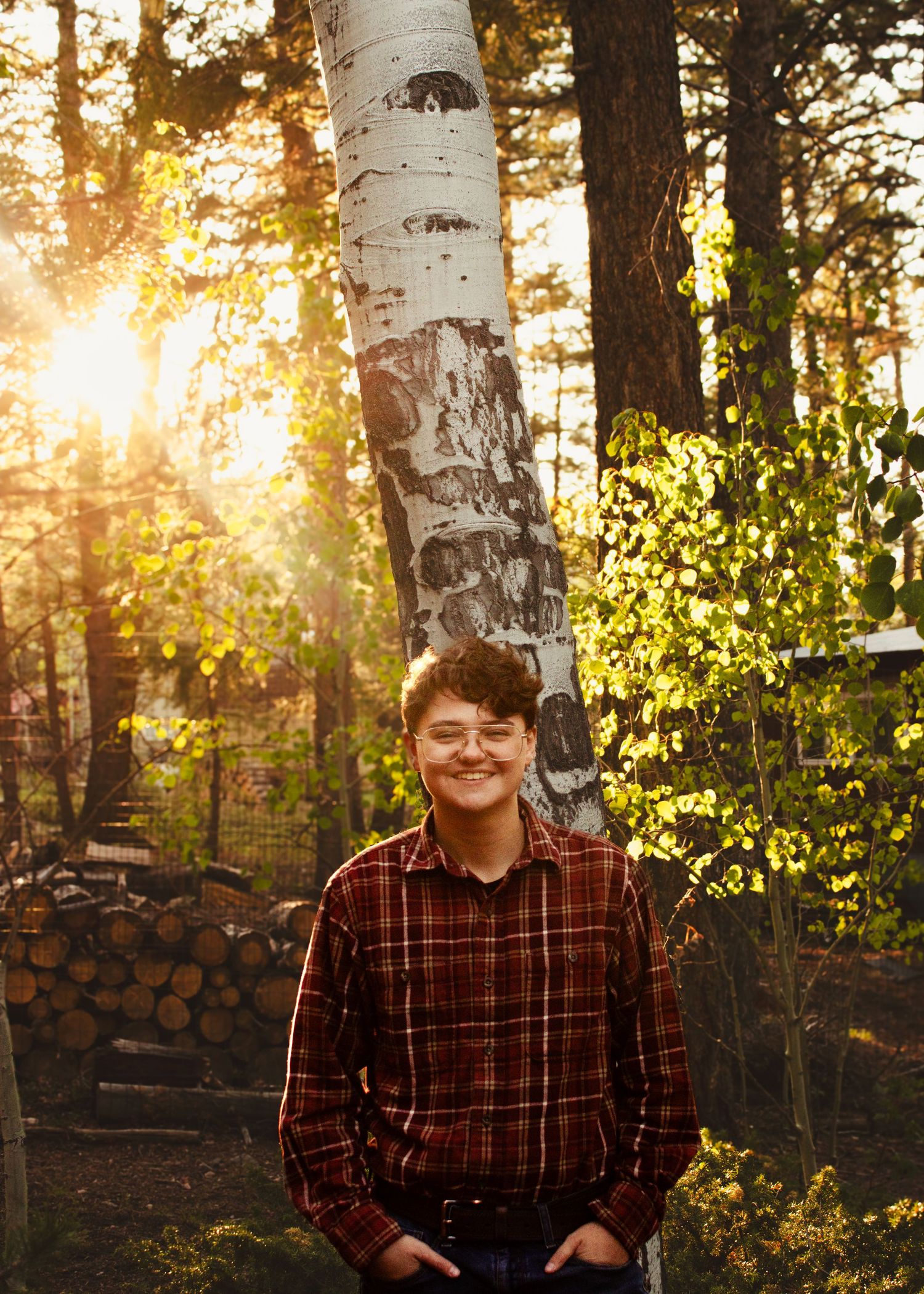 Smiling teen boy in plaid shirt standing by birch tree in sunlit forest.