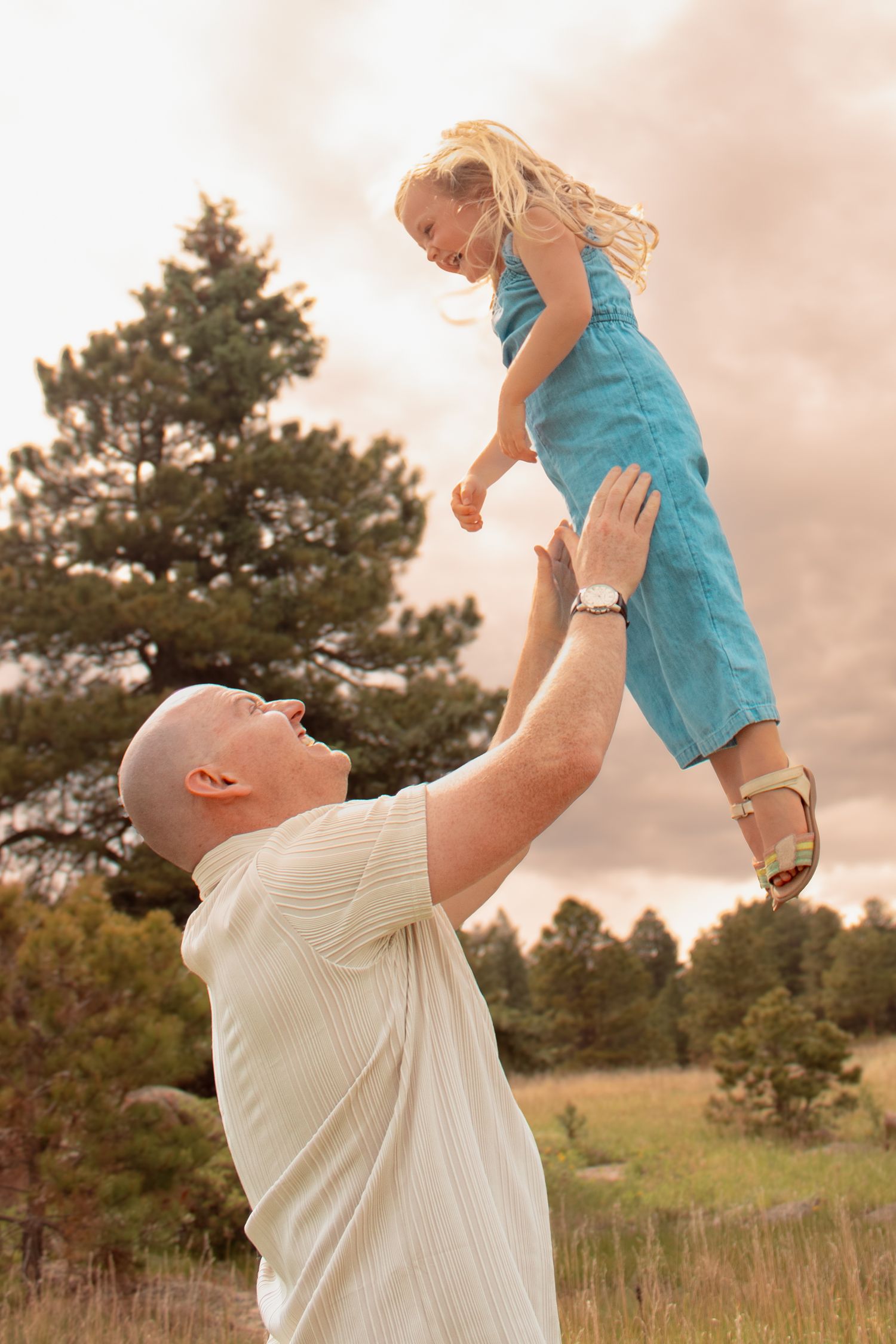 A bald man joyfully tosses a smiling blonde girl in a blue dress into the air outdoors.