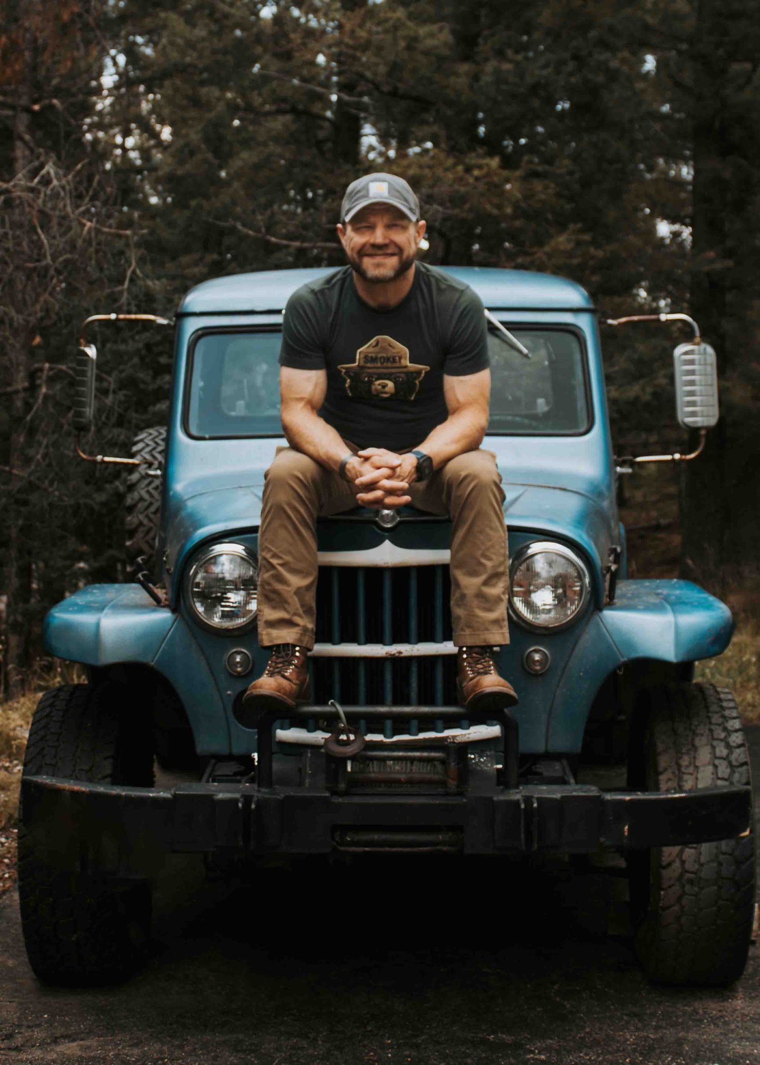 Man sitting on hood of vintage blue Jeep truck on forest dirt road, wearing gray cap and brown pants.