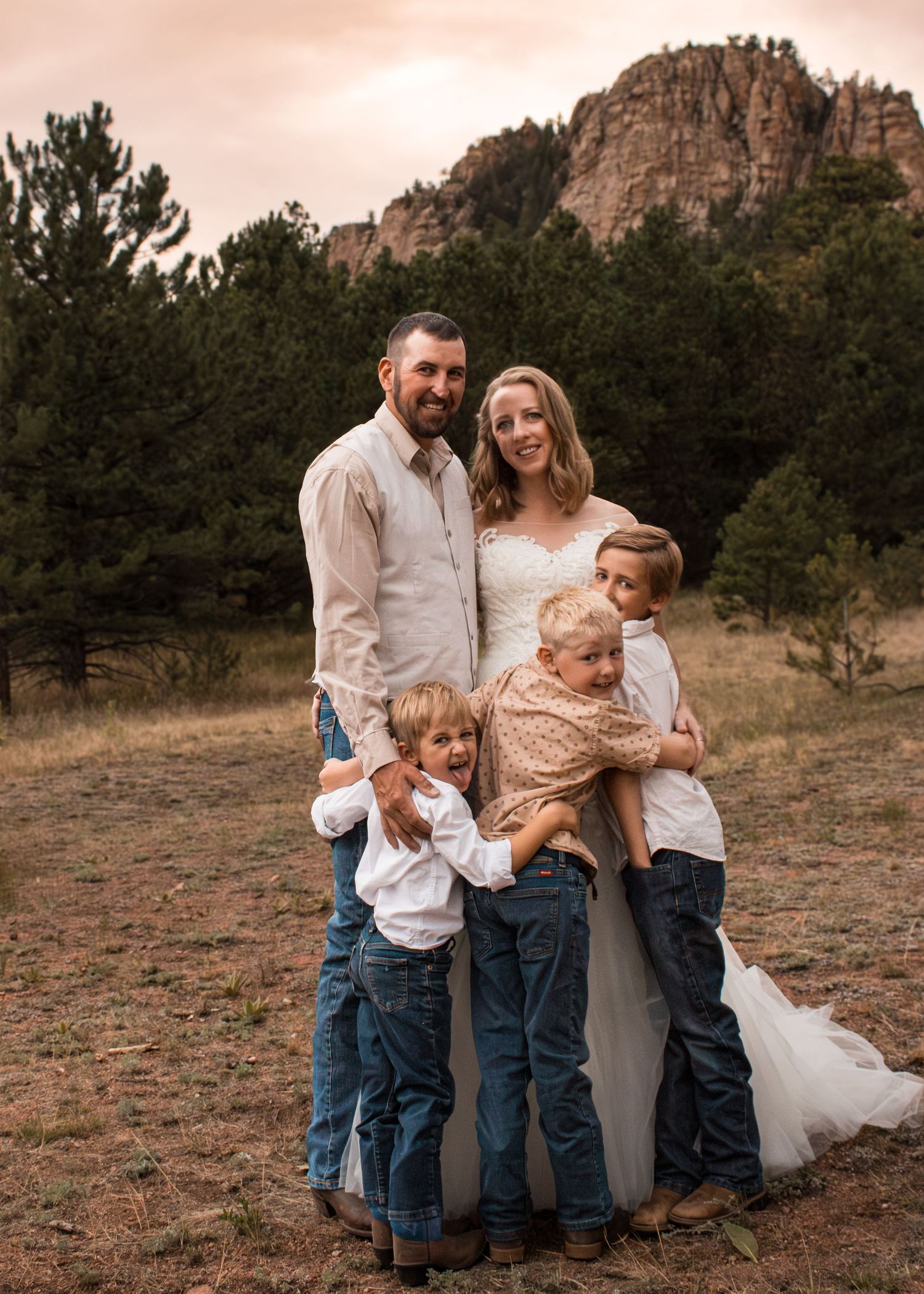 Family of five poses in rustic mountain meadow, parents in white and denim with three young boys, rocky peaks behind them.
