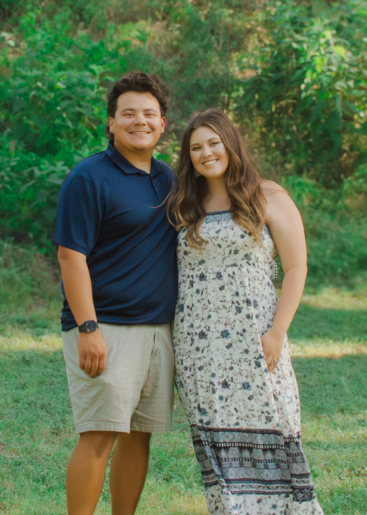 A young couple smiles outdoors in a green park, man in navy polo and khaki shorts, woman in floral sundress.