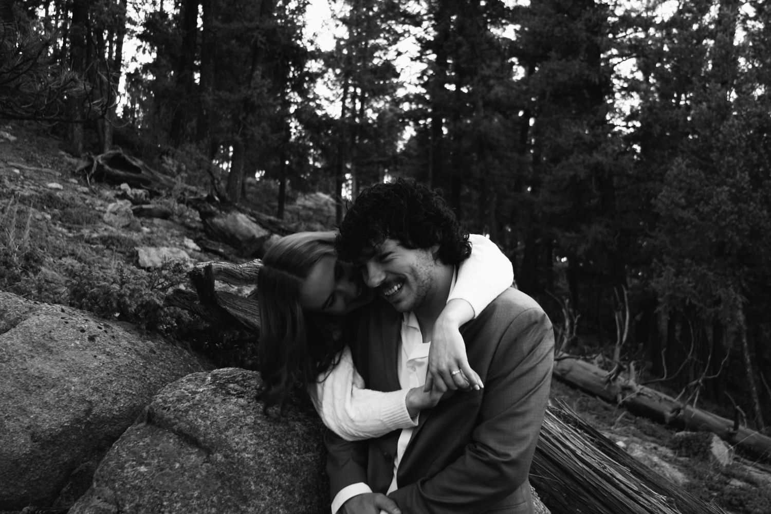 Black and white photo of a laughing couple sharing a playful piggyback moment on rocky forest terrain.