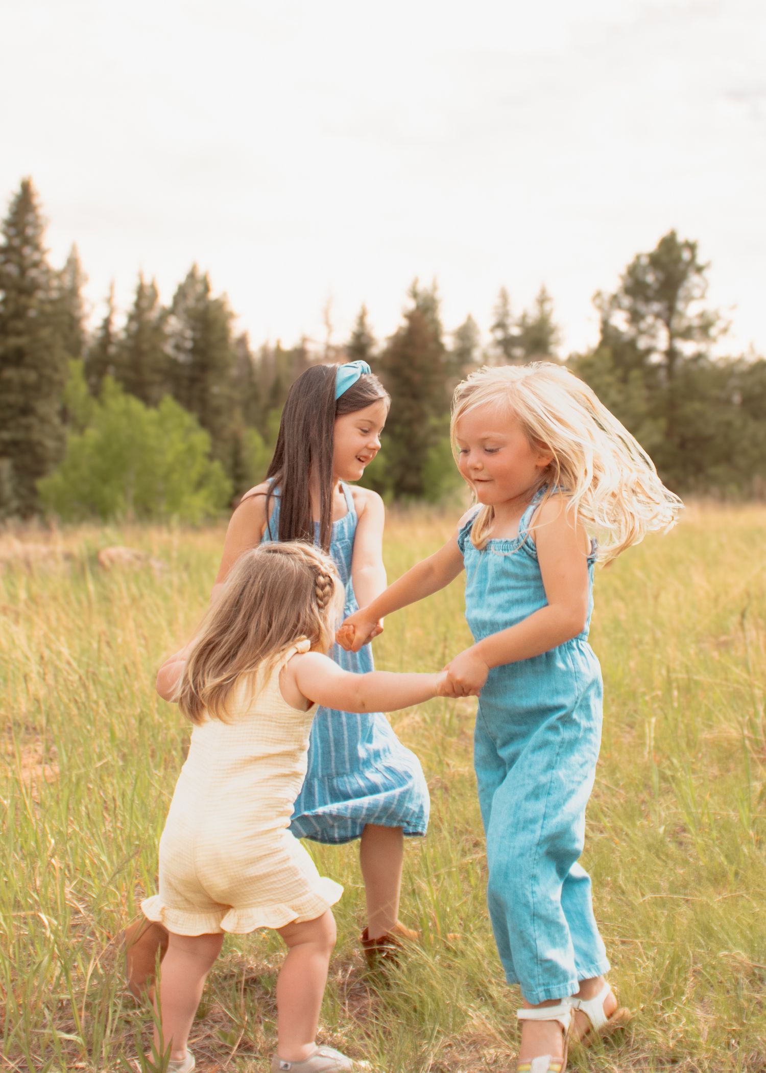 Three young girls hold hands and dance together in a sunny meadow surrounded by tall grass and pine trees.