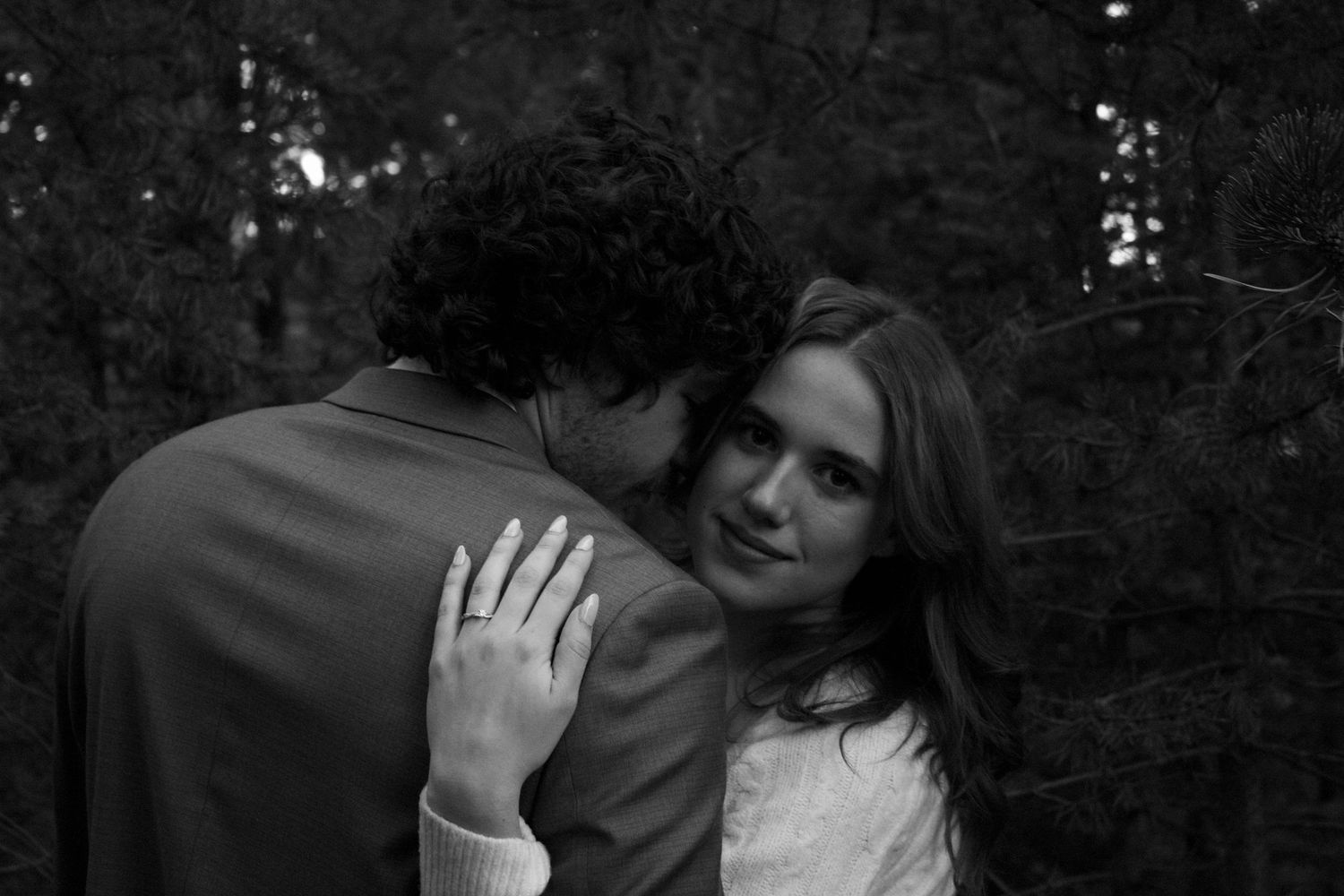 Intimate black and white portrait of a couple in the forest, woman smiling with engagement ring visible.