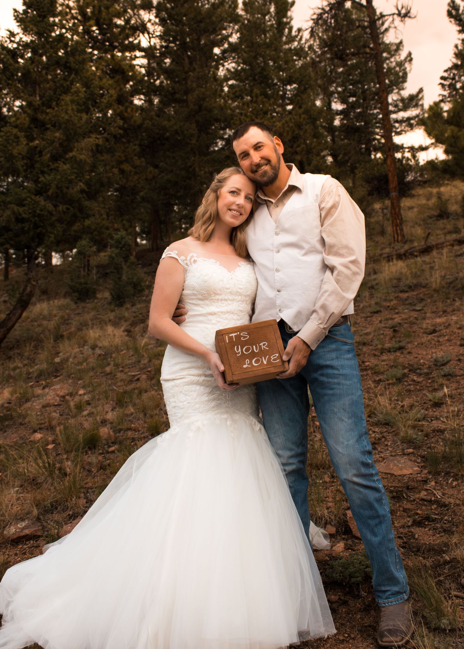A bride in a white mermaid gown and groom in jeans pose together holding a wooden sign in a forest setting.