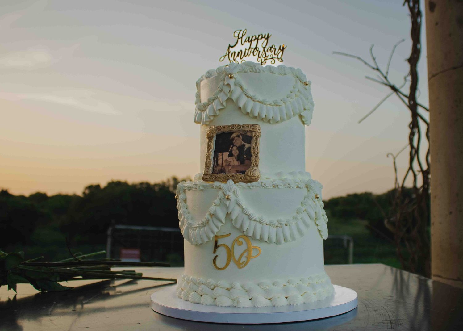 Elegant three-tier white anniversary cake with gold topper, draped frosting, photo, and 50th anniversary number decoration.