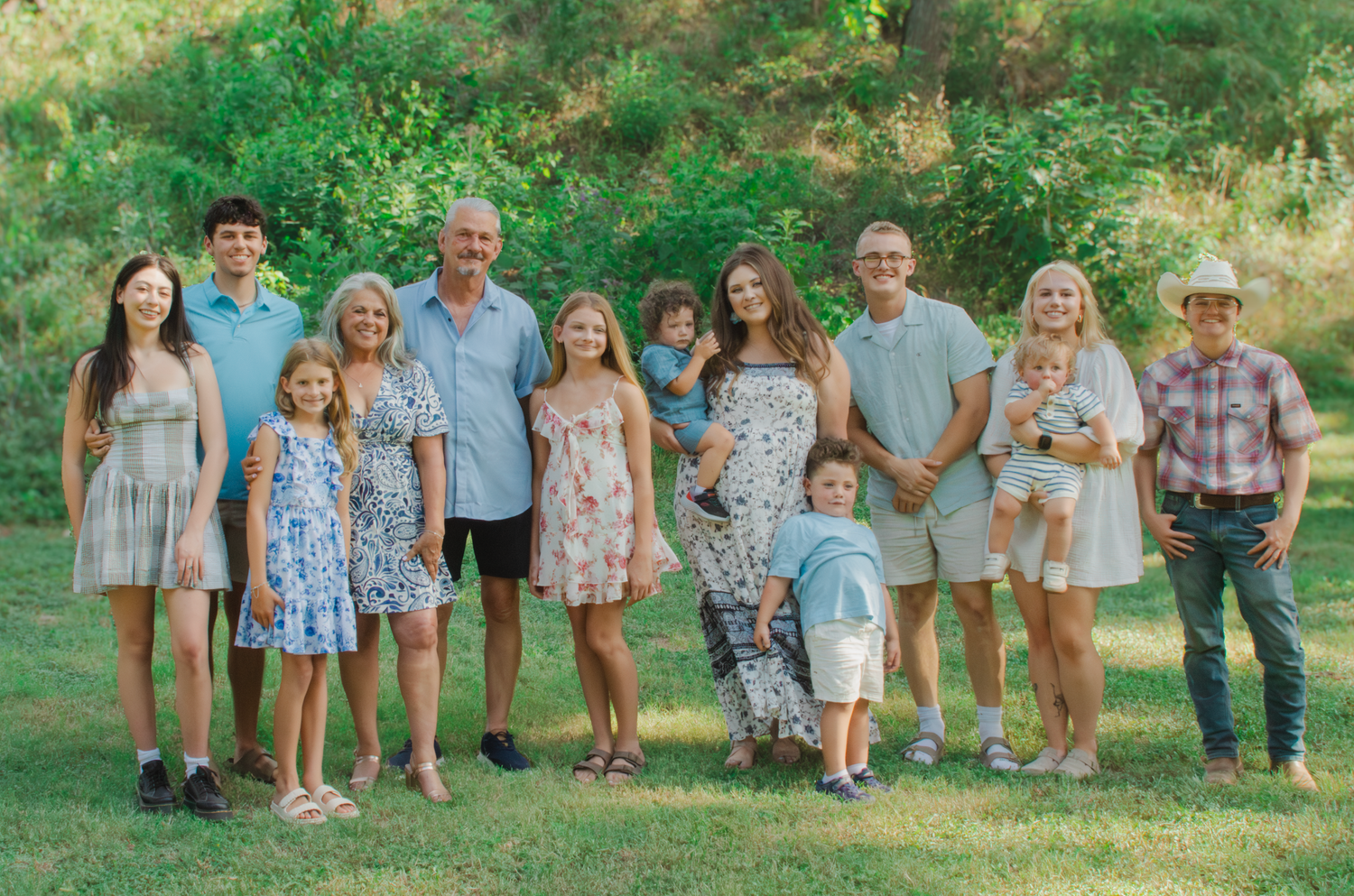 Large family group portrait outdoors on green grass, smiling adults and children in coordinated casual outfits for a summer photo session.