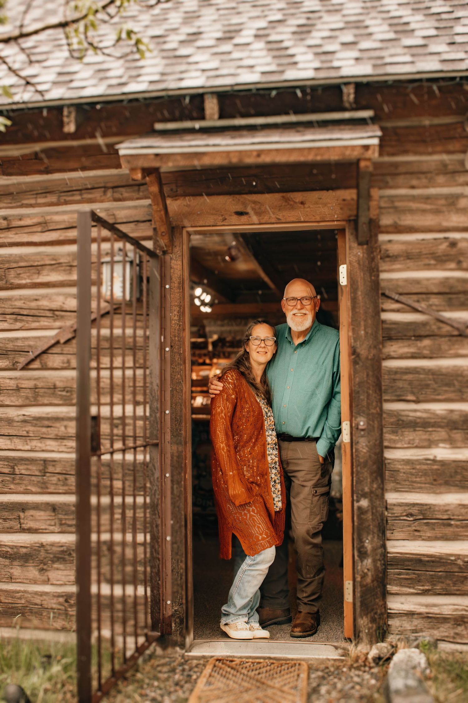 Smiling senior couple standing together in the doorway of a rustic wooden log cabin building.
