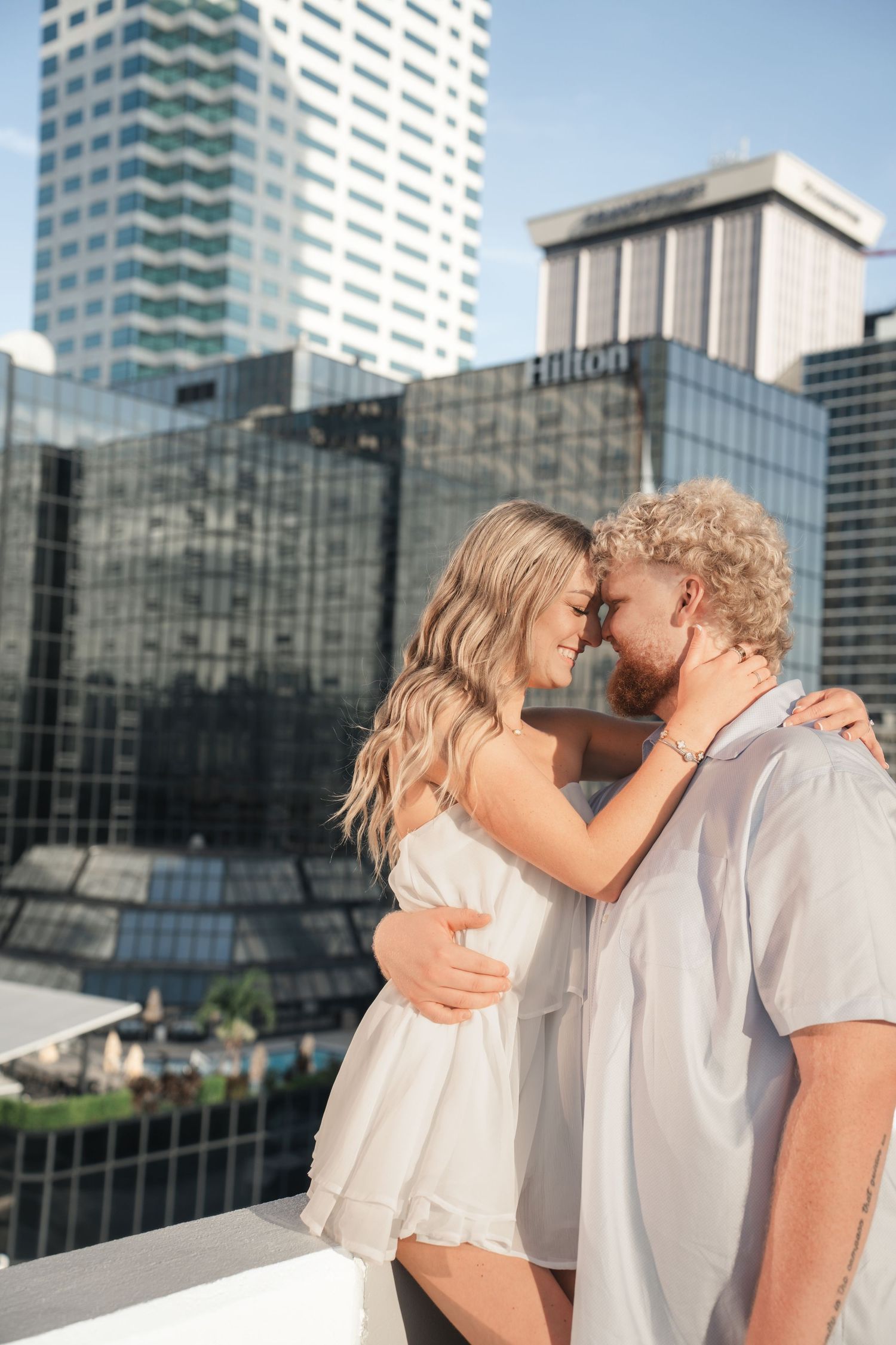 Couple embracing on rooftop with Hilton hotel and city skyscrapers in background on a sunny day.