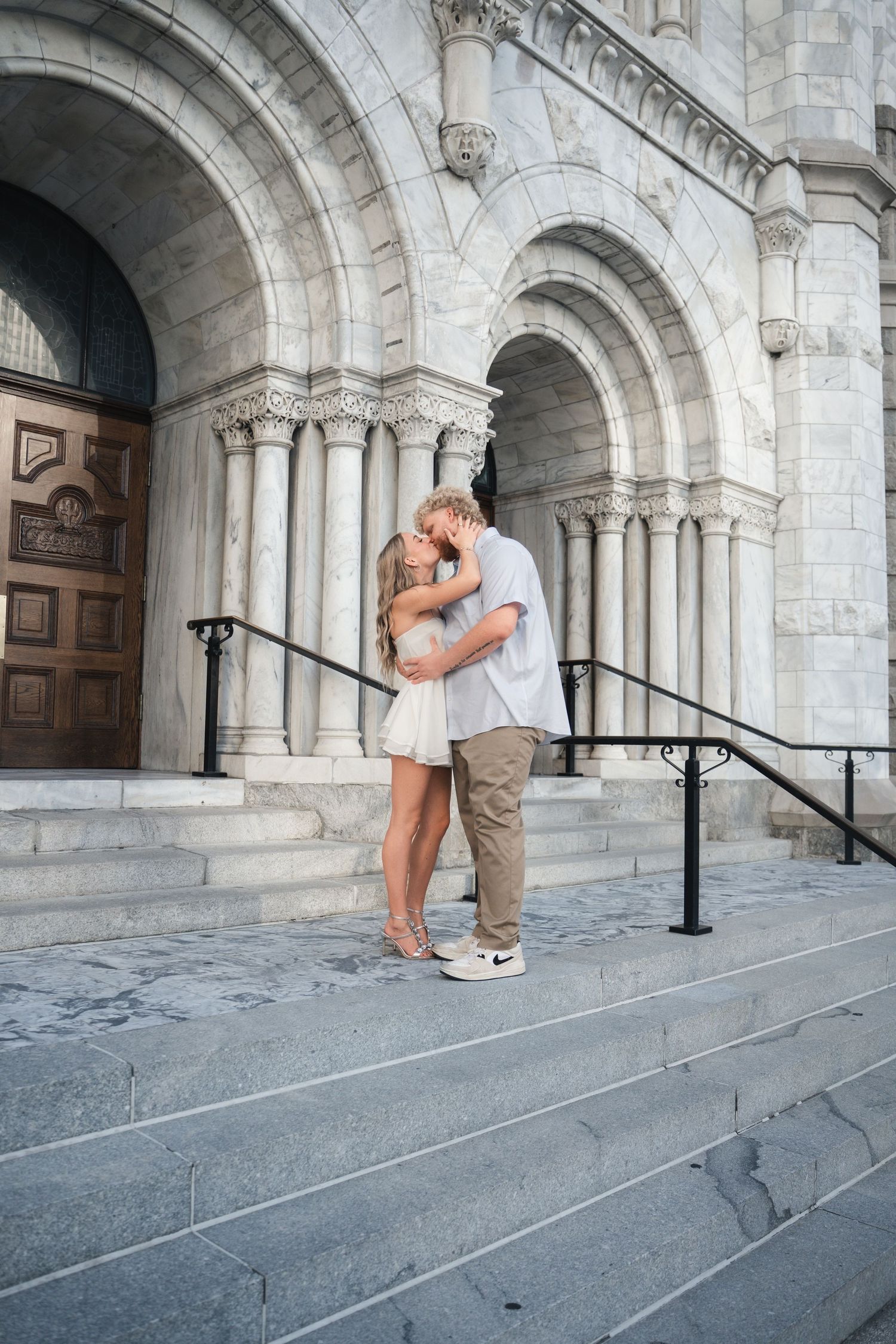 Couple sharing a romantic kiss on the steps of a grand white stone cathedral with ornate arches.