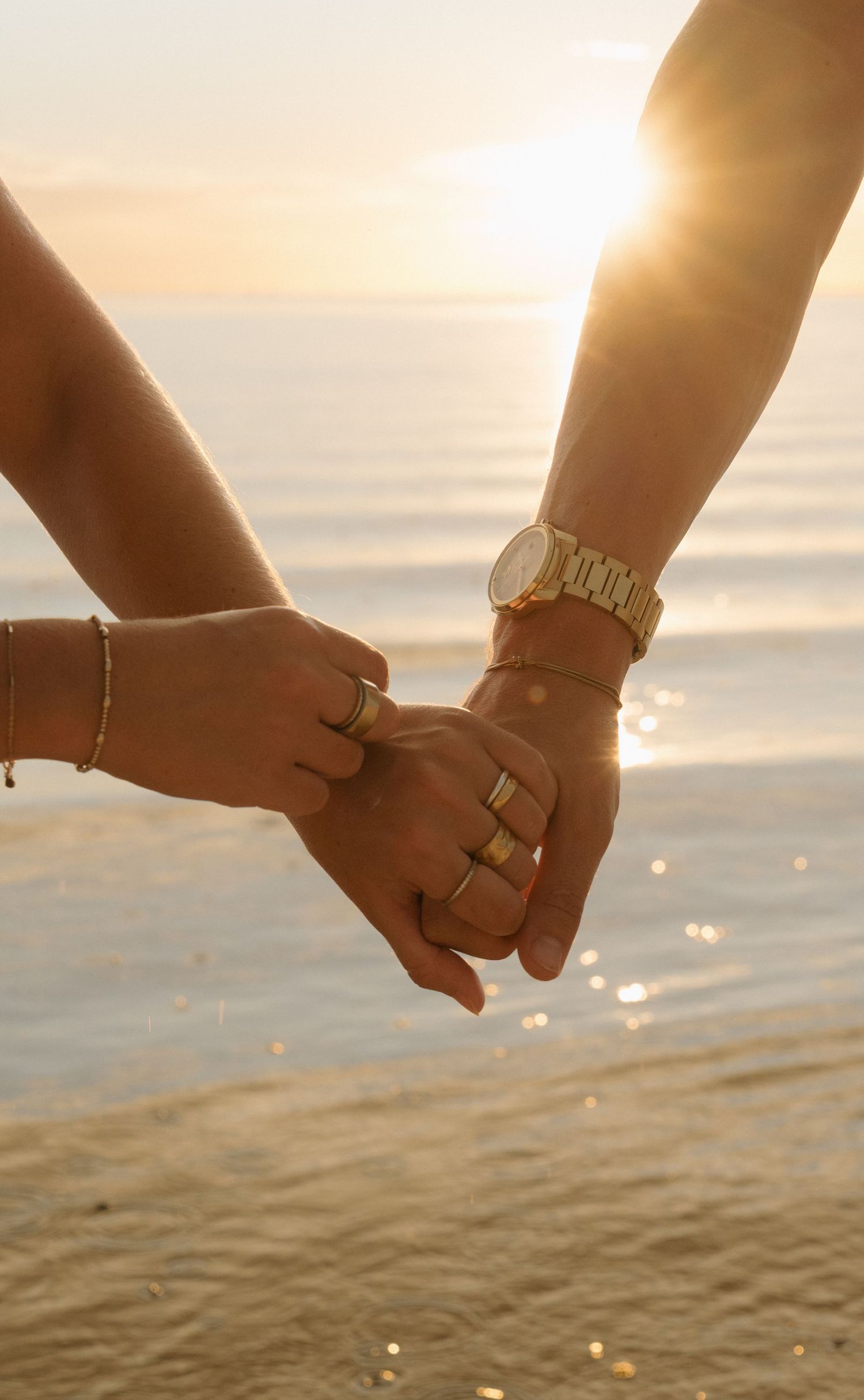 Couple holding hands at sunset on a beach, wearing gold rings, bracelets, and a gold watch, with ocean in background.