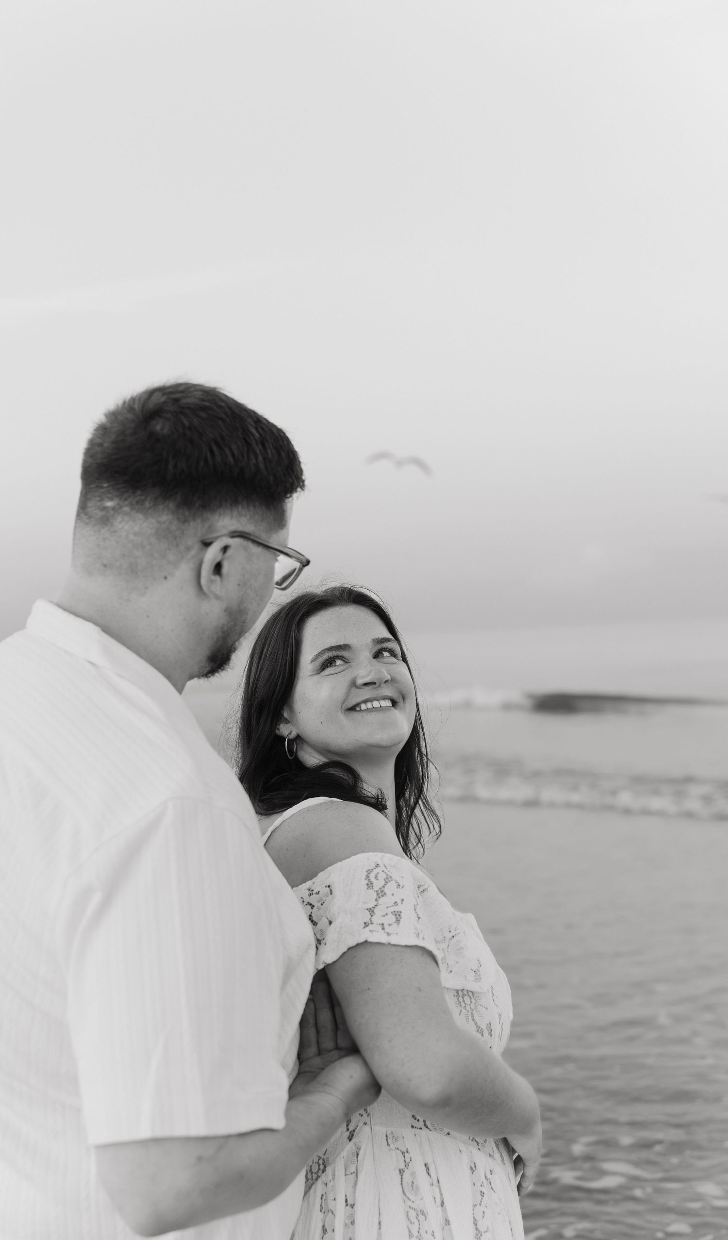 Black and white photo of a smiling couple embracing on a beach, woman in off-shoulder dress gazing up lovingly.