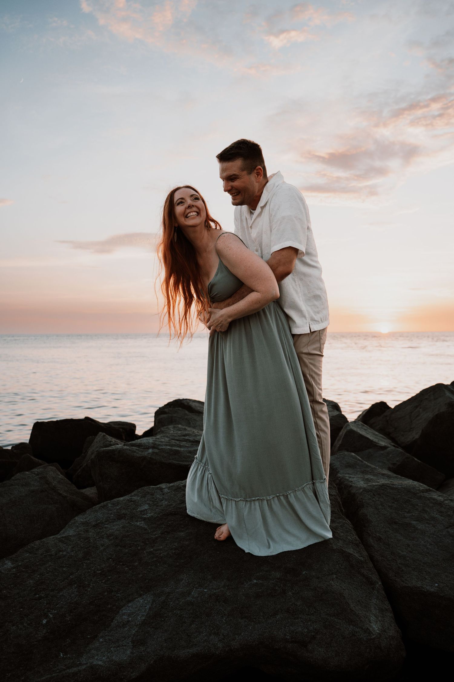 Couple embracing on rocky shore at sunset, woman in sage green maxi dress laughing joyfully.