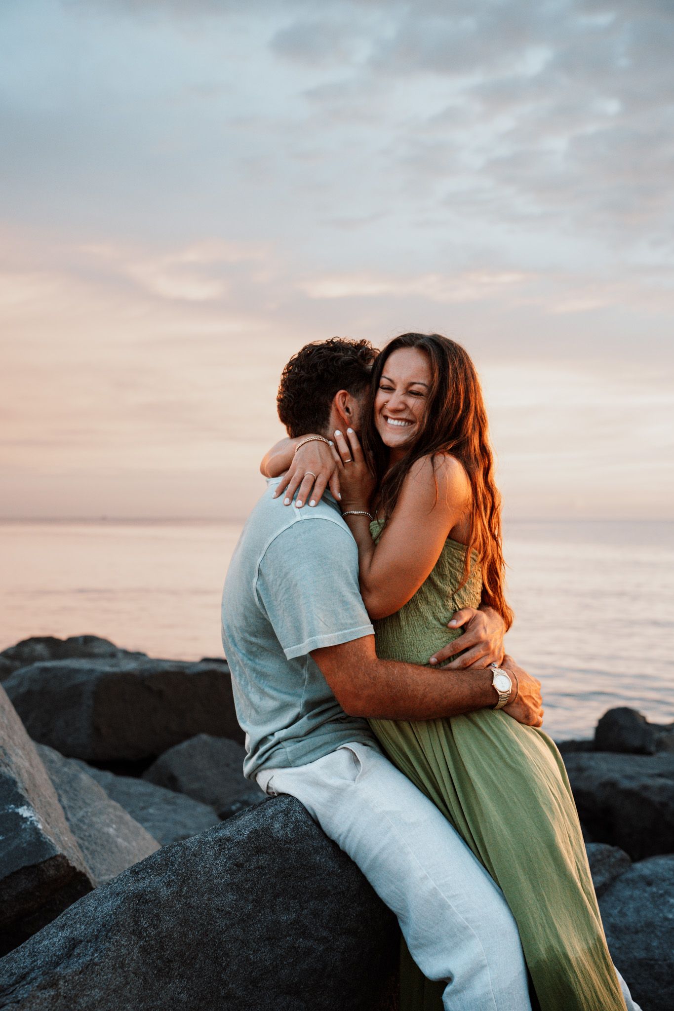 Couple embracing on rocks at sunset by the ocean, woman in green dress laughing joyfully.