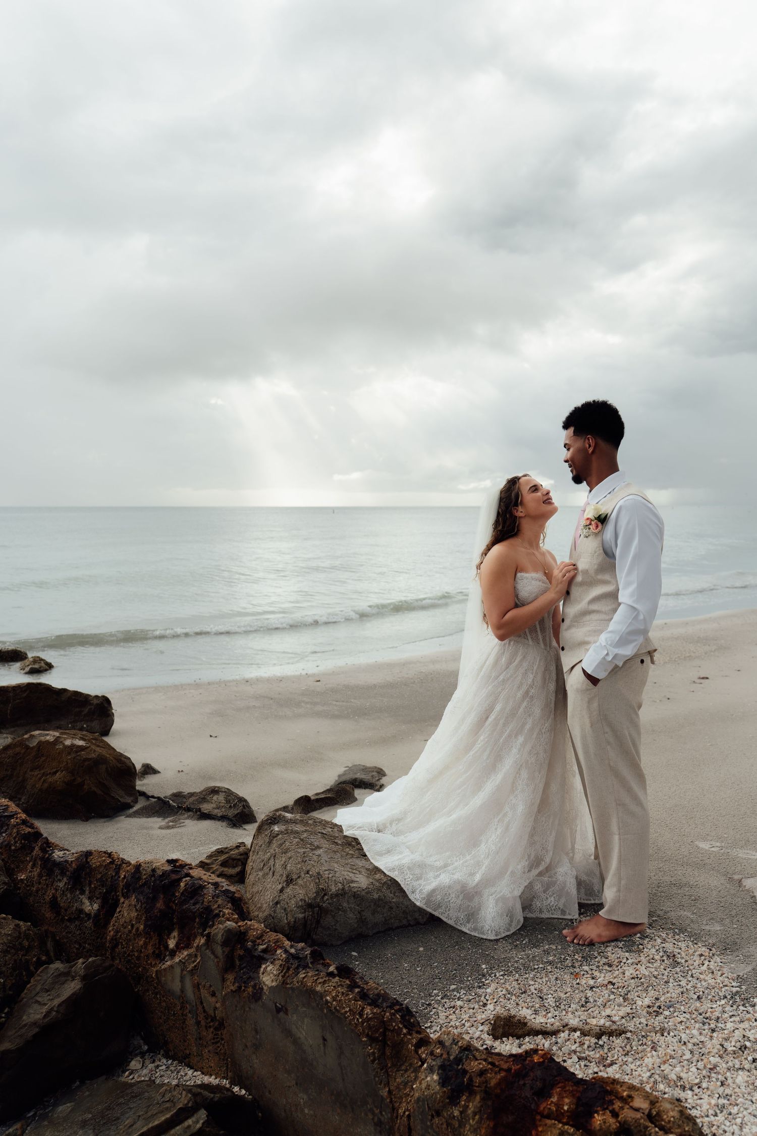Bride and groom embrace on rocky beach with ocean waves and dramatic cloudy sky behind them.