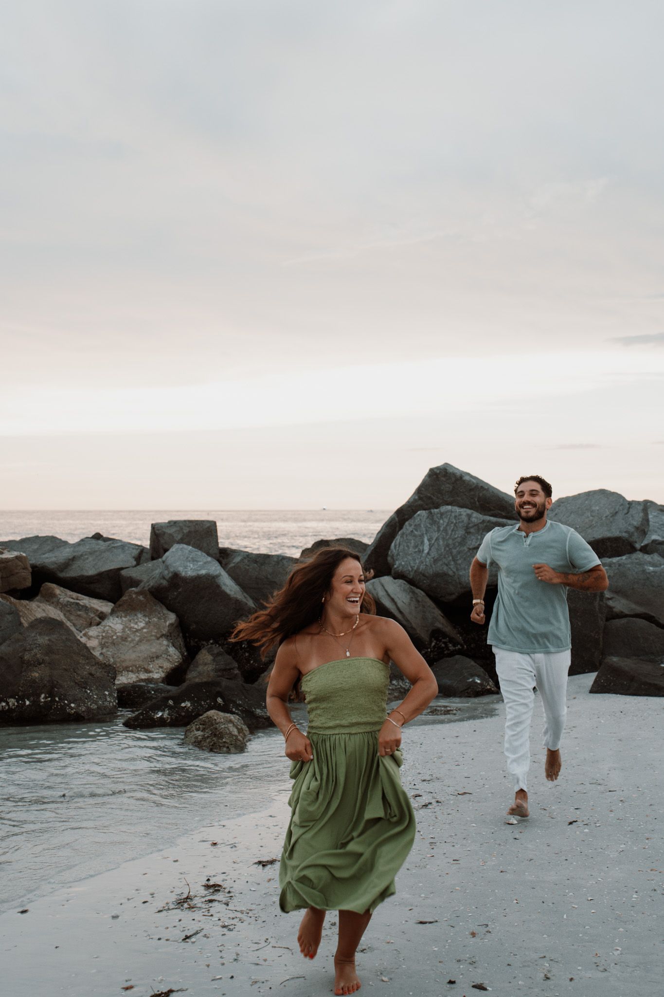 A joyful couple runs along a rocky beach at sunset, woman in green dress, man in blue shirt and white pants.