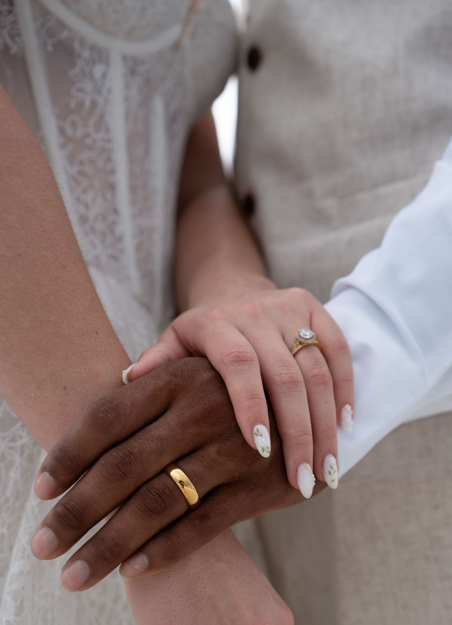 Close-up of interracial couple holding hands showing gold wedding bands and diamond engagement ring with white floral nail art.