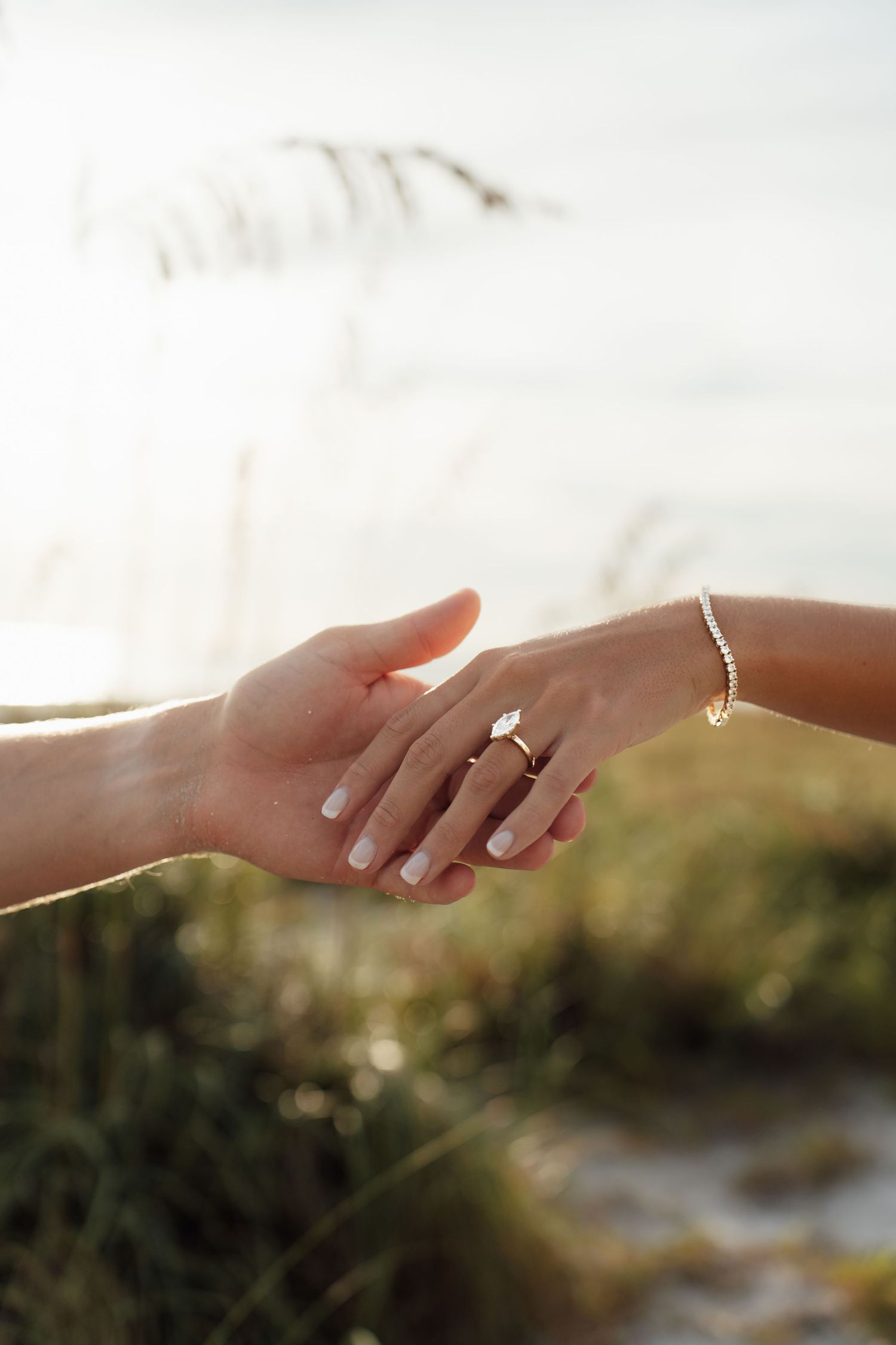 Two hands holding gently outdoors, woman wearing a gold diamond engagement ring and tennis bracelet.