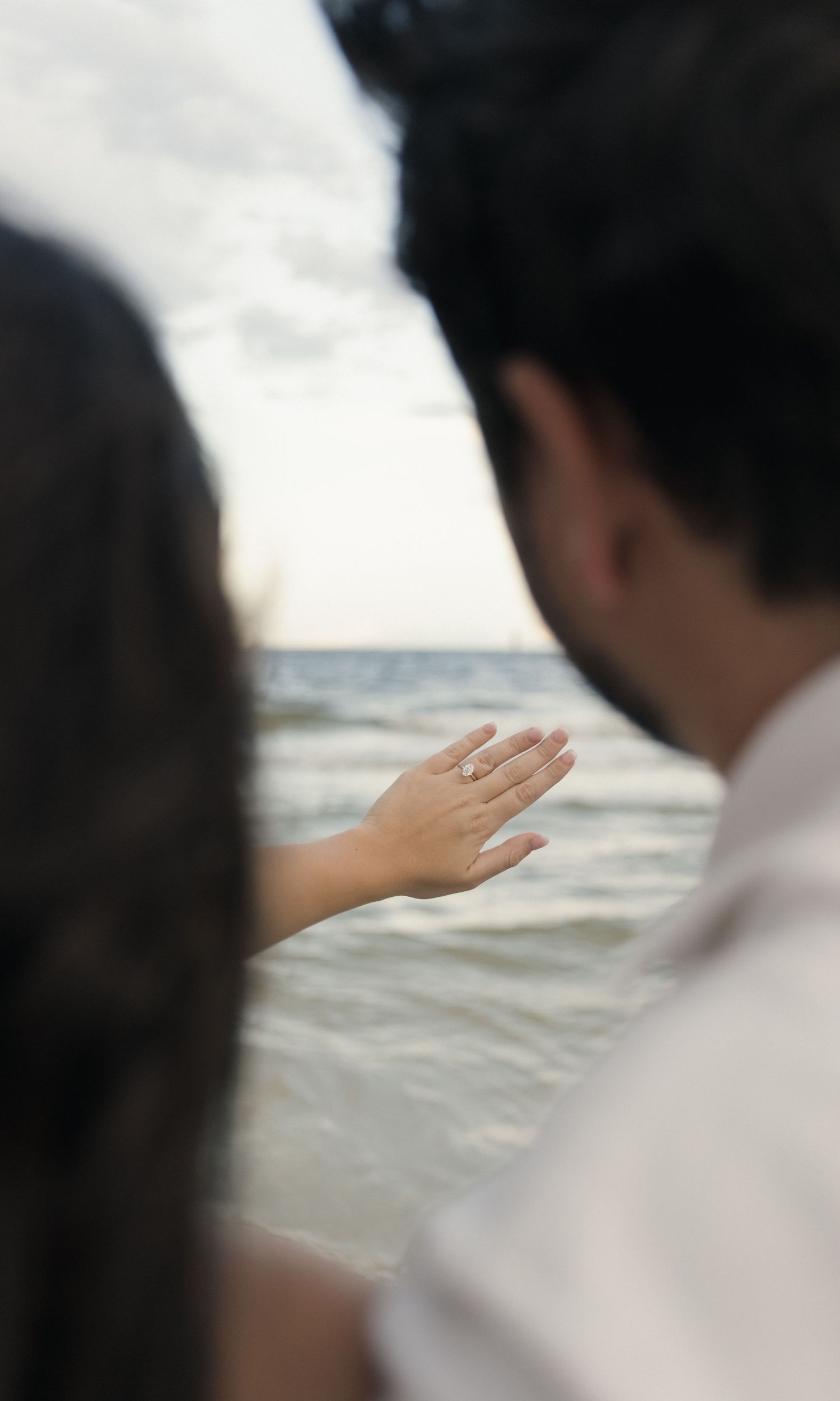 Couple at beach with woman showing off engagement ring while looking at ocean waves.