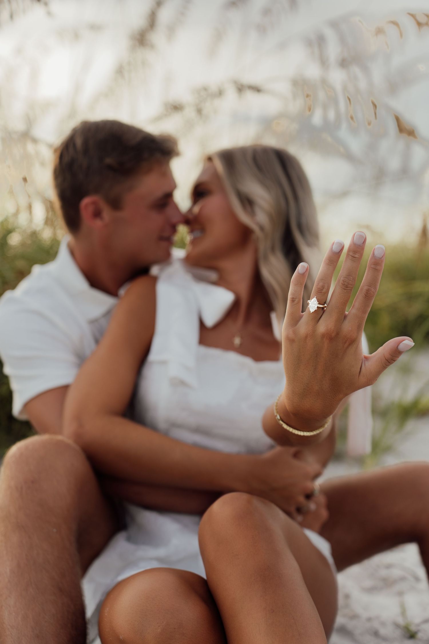 Couple in white outfits sharing a romantic moment while showing off an engagement ring on the beach.