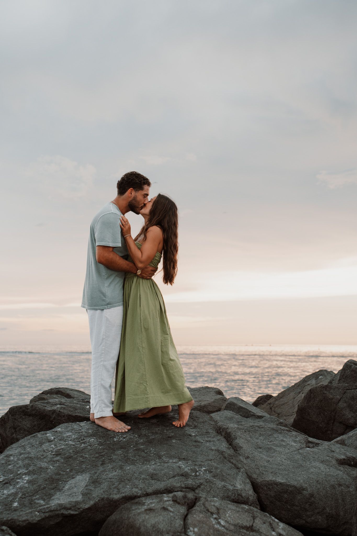 Couple kissing on rocky shore at sunset, woman in green dress and man in casual attire embracing by the ocean.