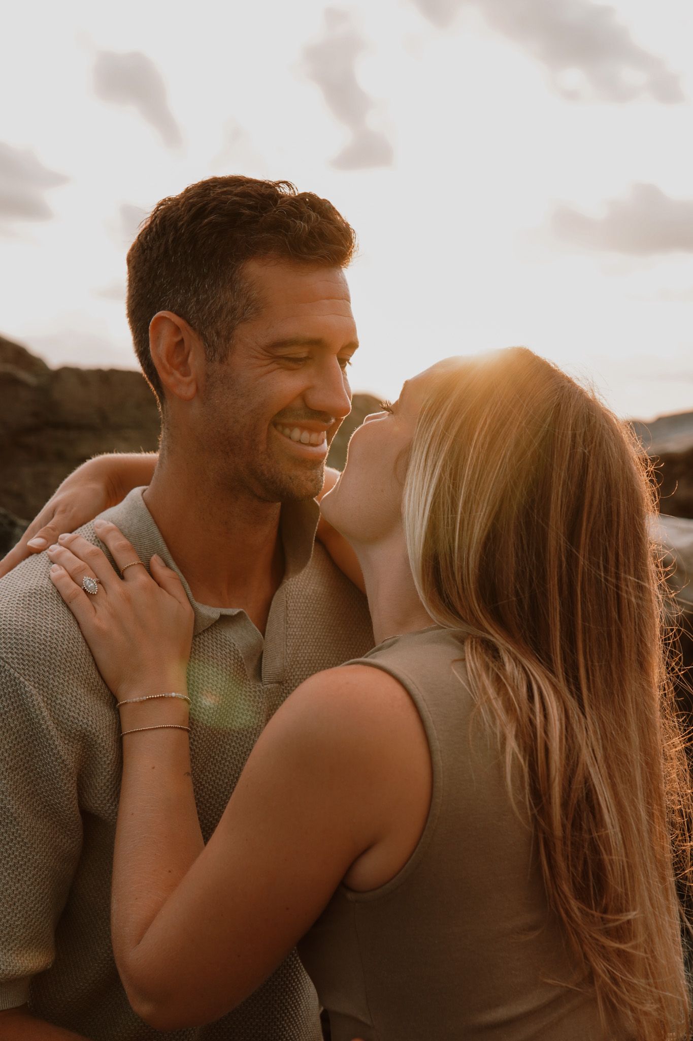A smiling couple shares a romantic golden hour moment outdoors, with the woman embracing the man as warm sunlight glows behind them.
