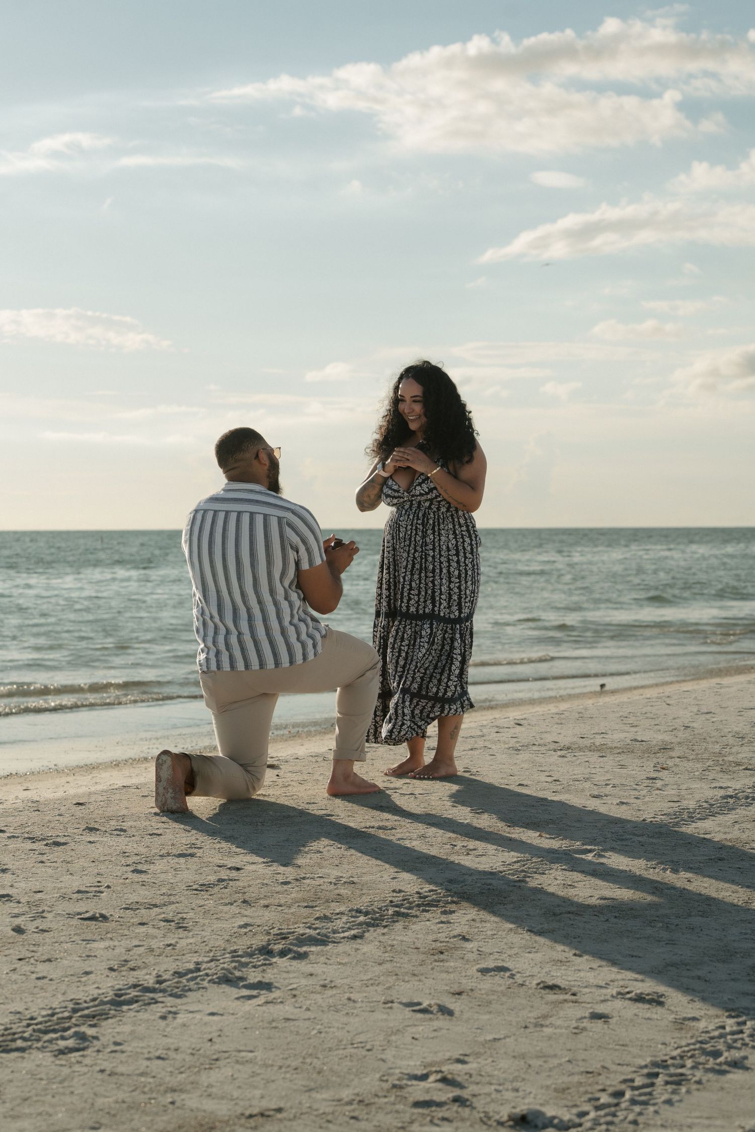 Man proposes on one knee to woman on a sandy beach at sunset with ocean in the background.