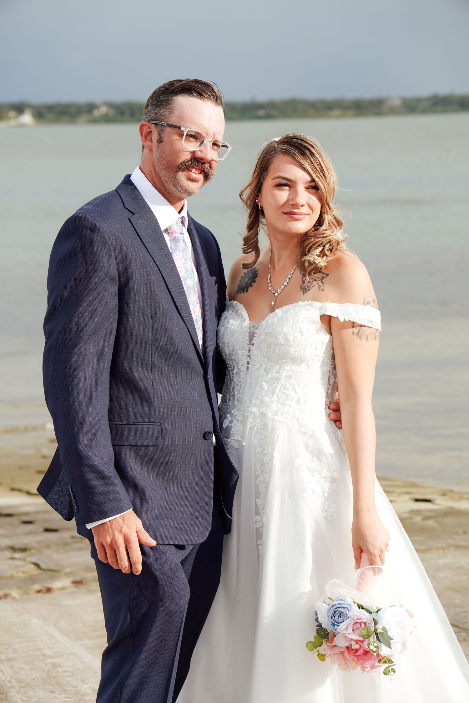 A bride and groom pose together on a beach, she in a white off-shoulder gown holding a bouquet, he in a navy suit.
