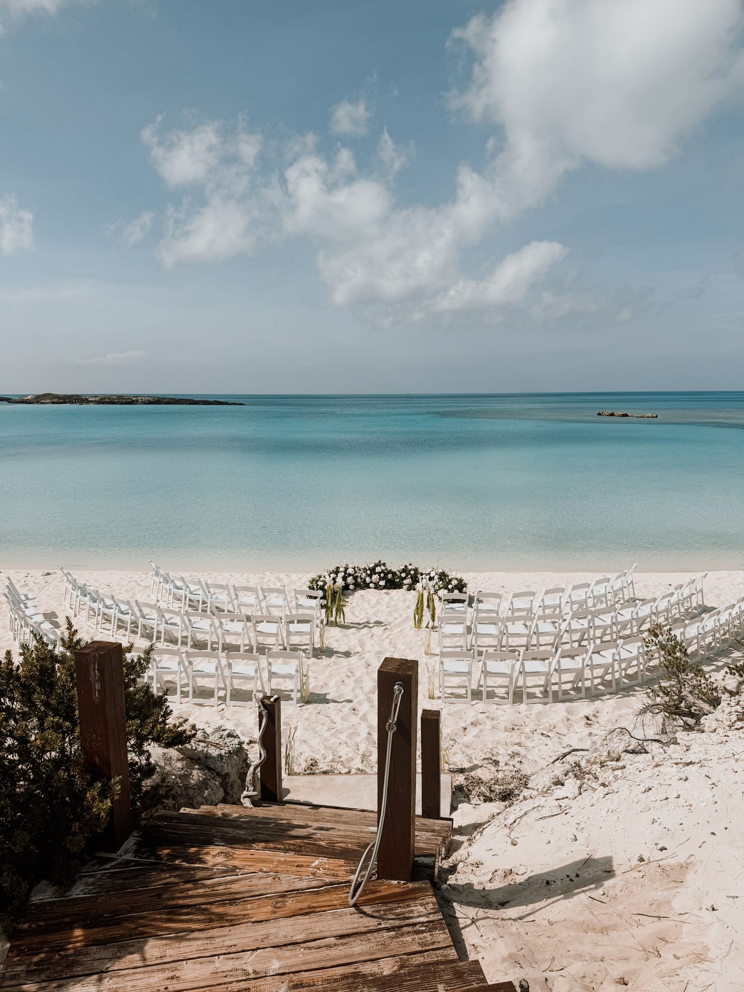 Beachfront wedding ceremony setup with white chairs and floral arch on white sand beach with turquoise water.