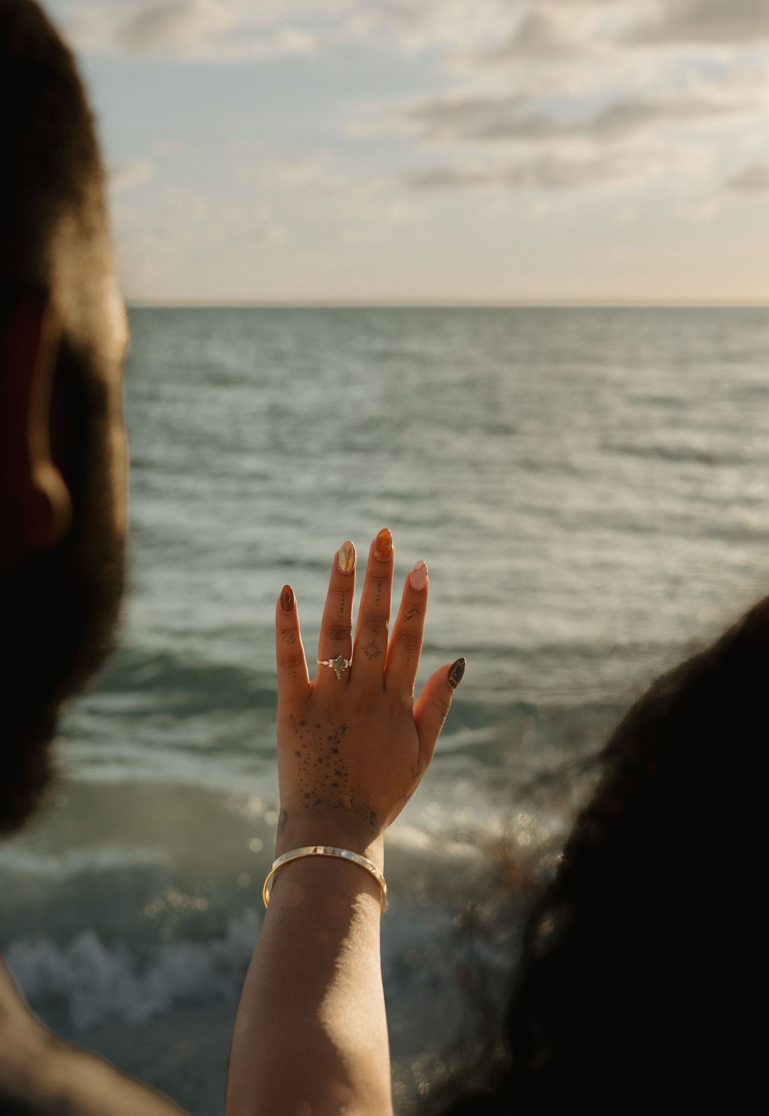 A woman displays her engagement ring and bracelet with the ocean in the background at sunset.