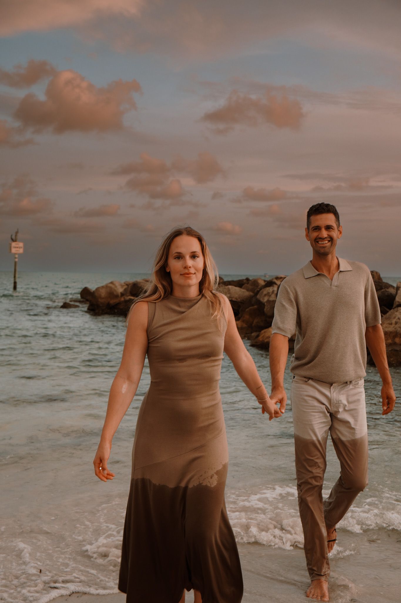 Couple holding hands walking on beach at sunset, woman in olive dress, man in casual attire, waves and rocks behind.