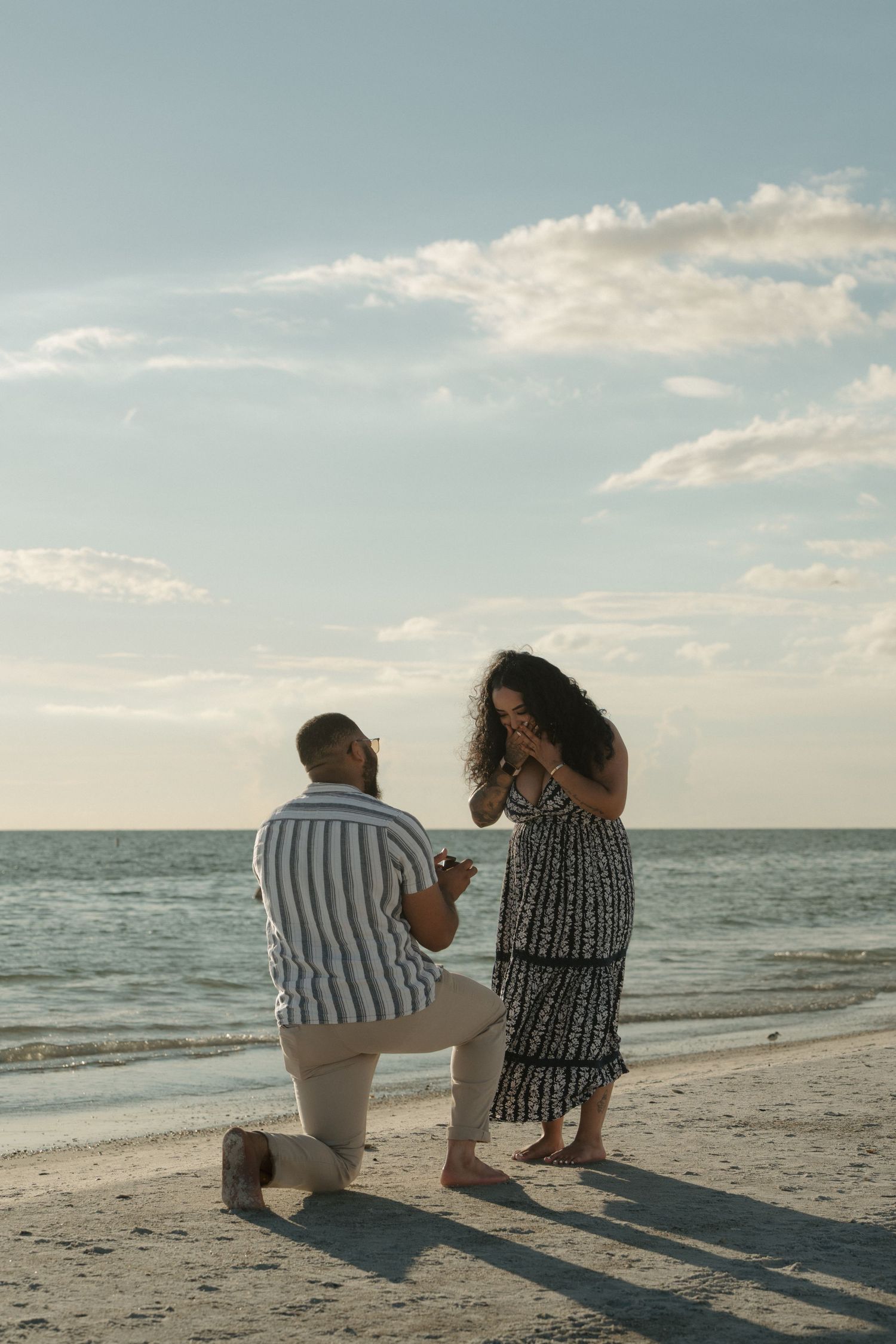 Man proposing on one knee to woman on a sandy beach at sunset, she covers her mouth in surprise.