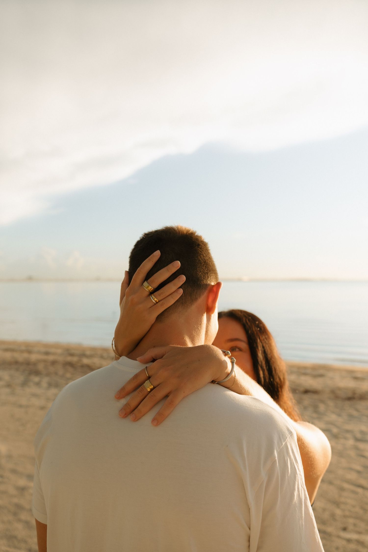 Couple embracing on a sandy beach at sunset, woman showing engagement ring while hugging man in white t-shirt.