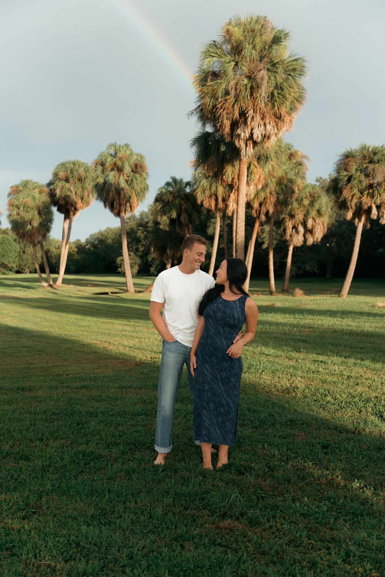 Couple standing on green grass with palm trees and rainbow in background on a sunny day.