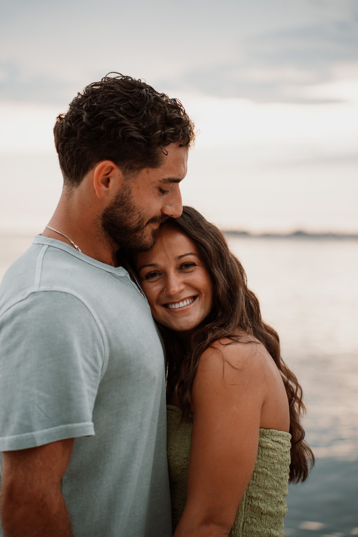A smiling couple embraces near the water at sunset, woman in green top leans against man in light blue tee.