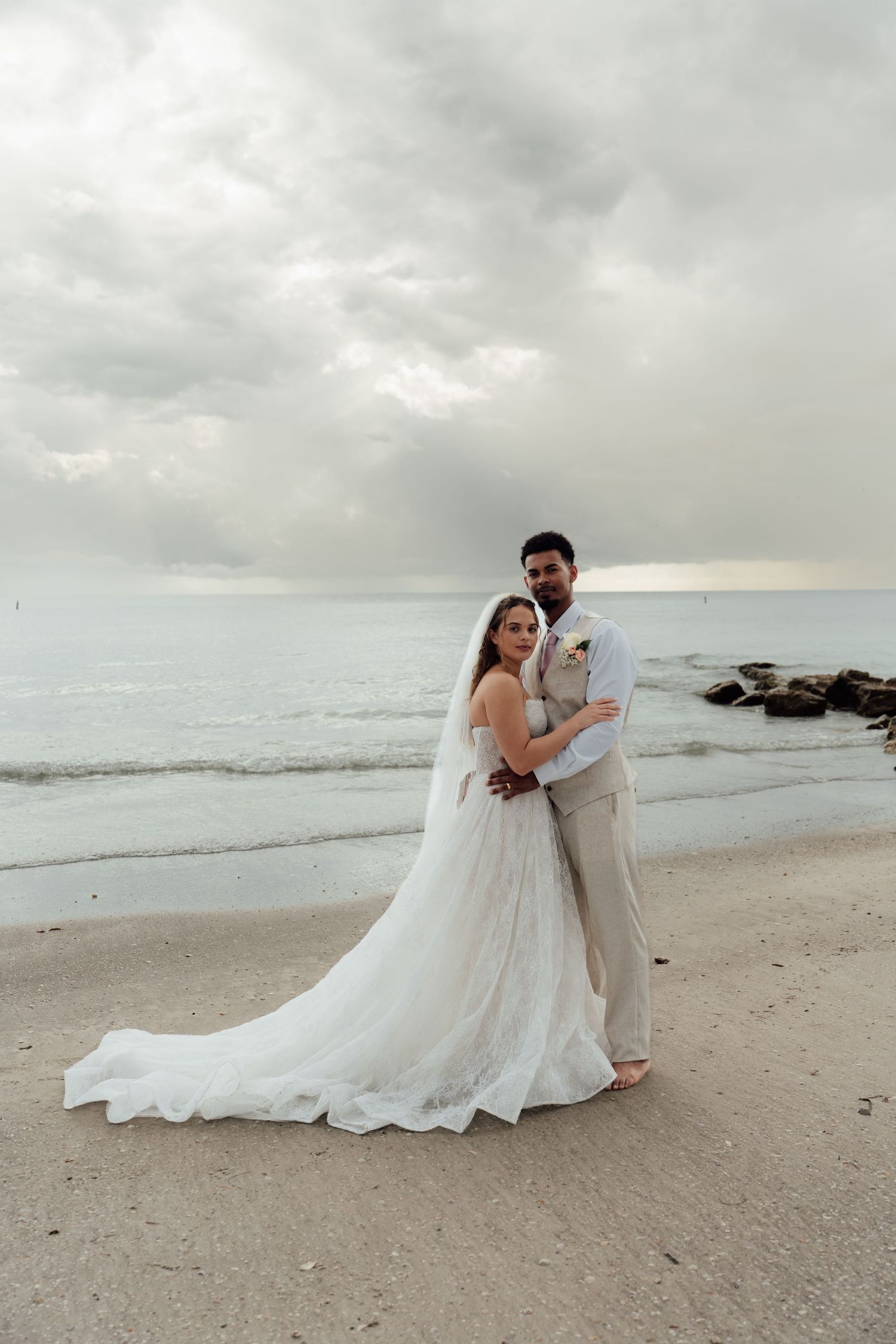 Bride and groom embracing on a sandy beach with ocean waves and cloudy sky backdrop on their wedding day.