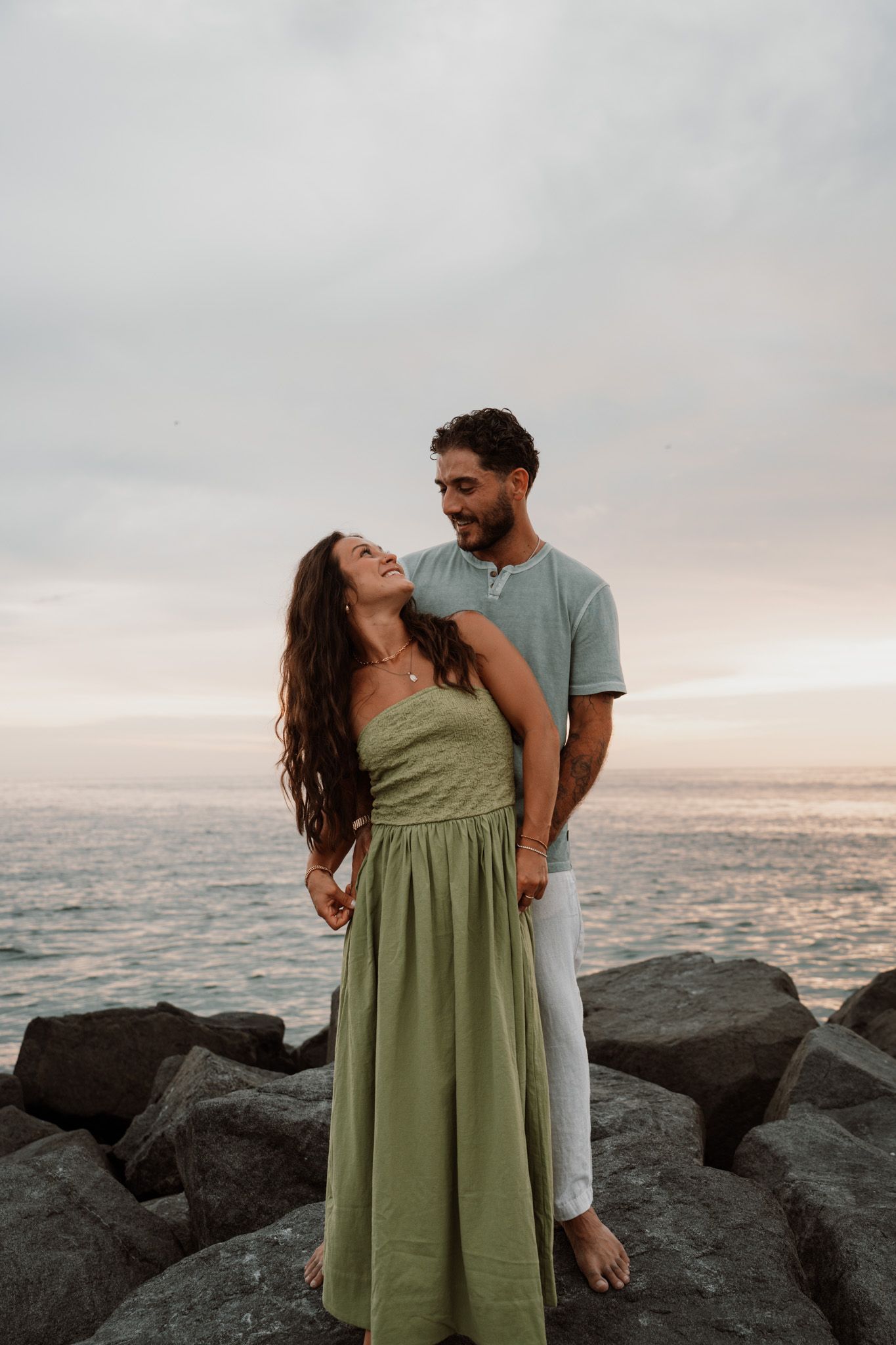 A romantic couple stands on beach rocks at sunset, woman in green maxi dress gazing up at her partner.
