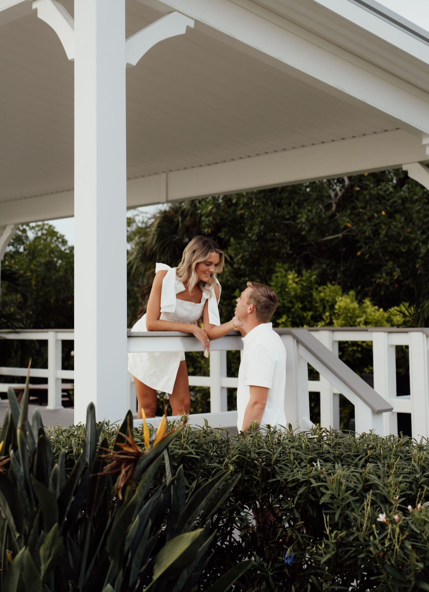 Couple in white outfits sharing a romantic moment on a white gazebo porch surrounded by tropical greenery.