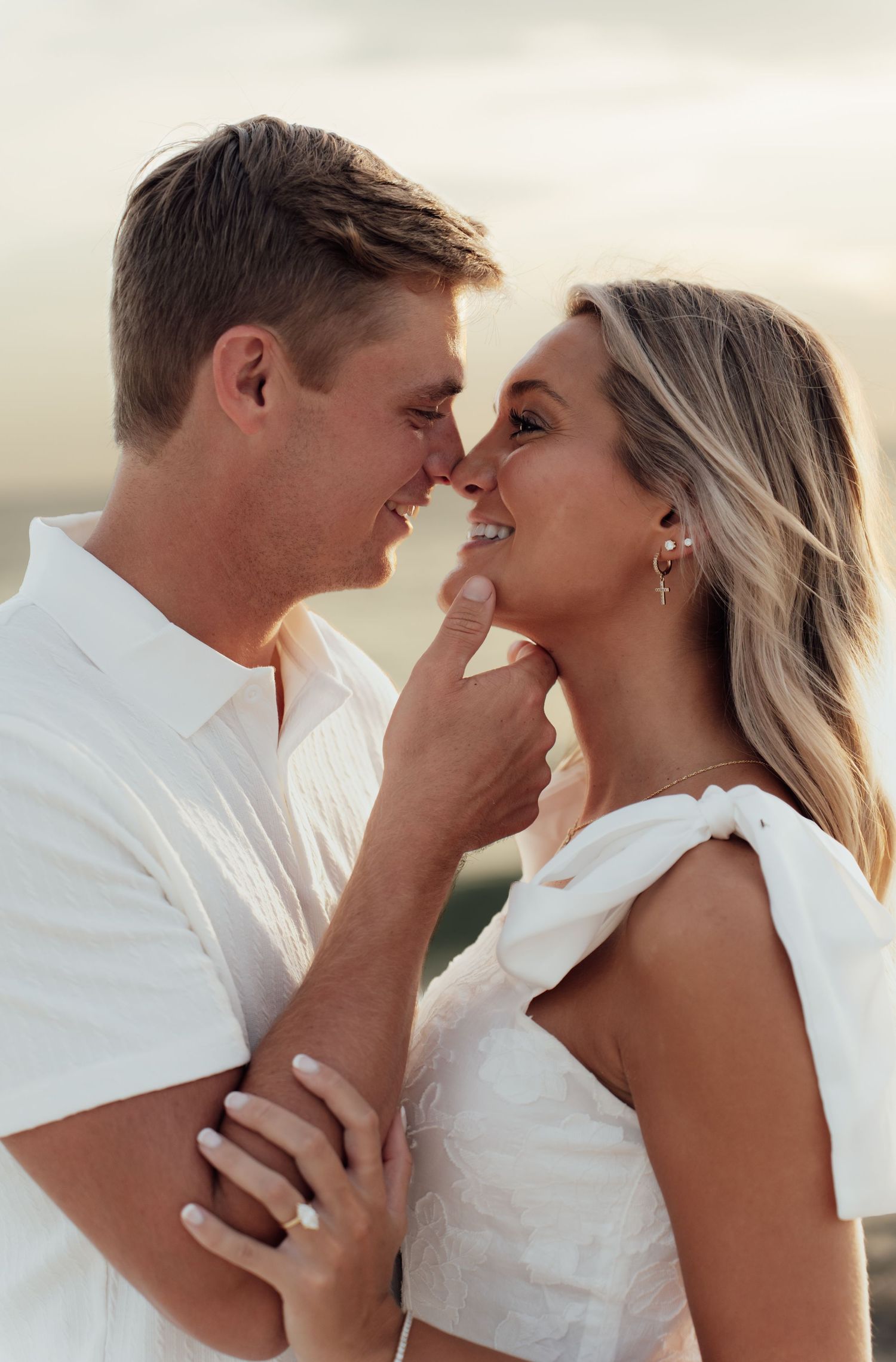 Couple in white outfits share a romantic moment, smiling close together outdoors at golden hour.