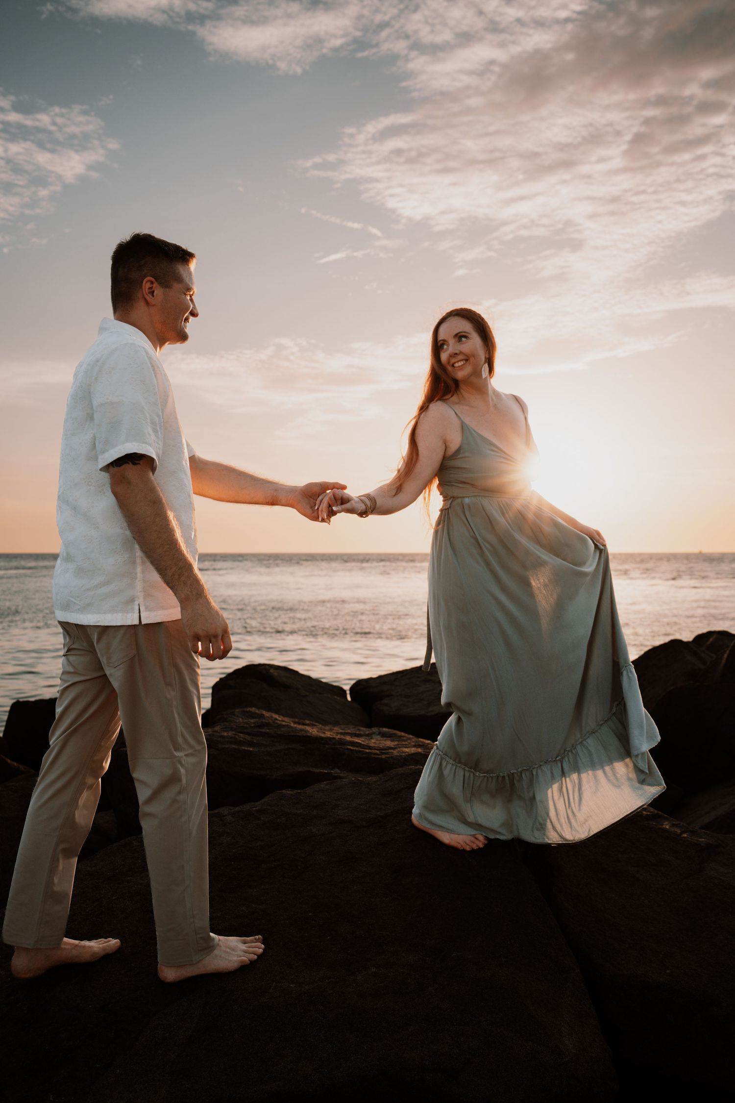 Couple holding hands on rocky beach at sunset, woman in flowing teal dress and man in white shirt, ocean backdrop.
