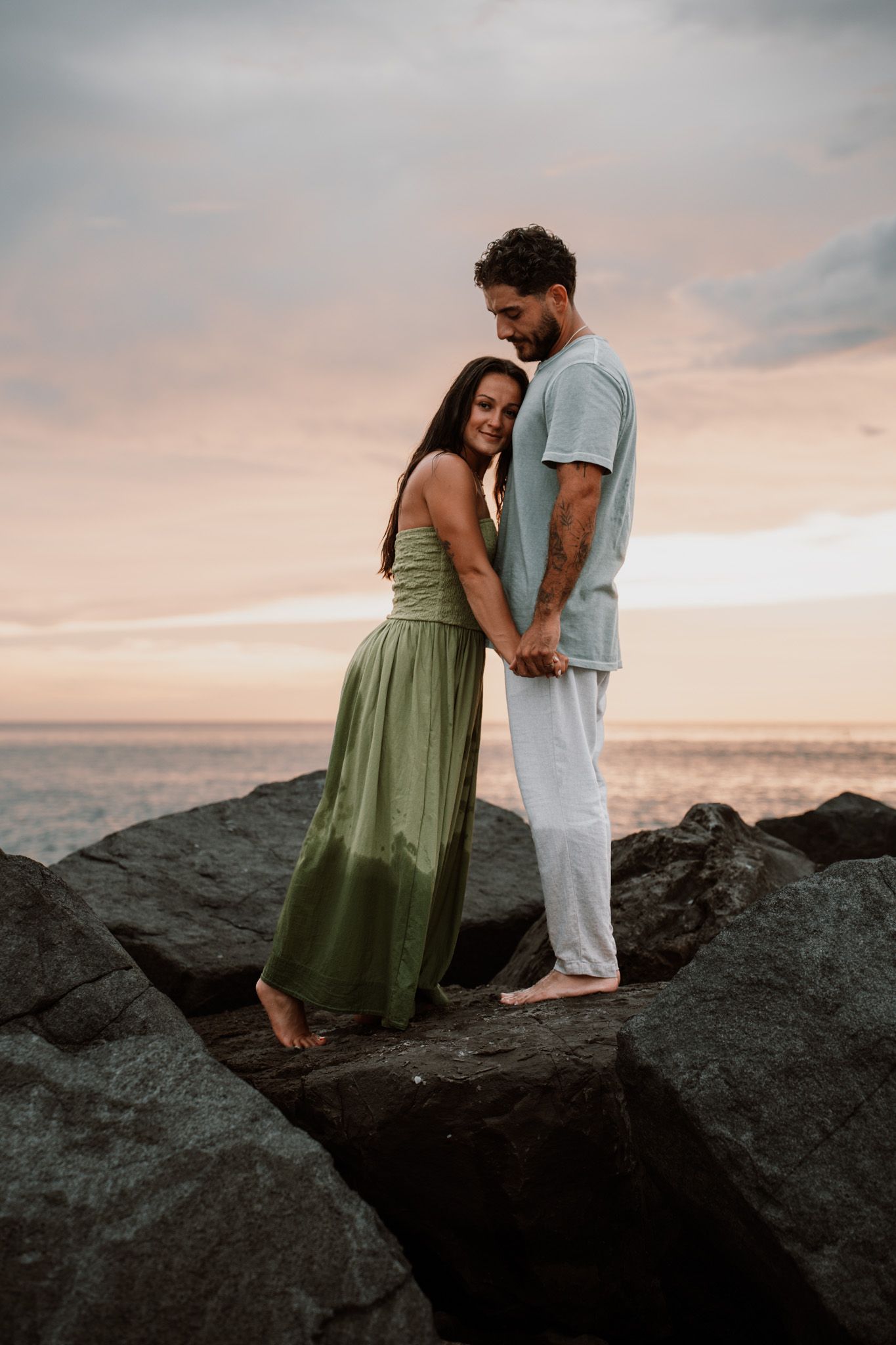 Couple holding hands on rocky shore at sunset, woman in green dress, man in light blue outfit.