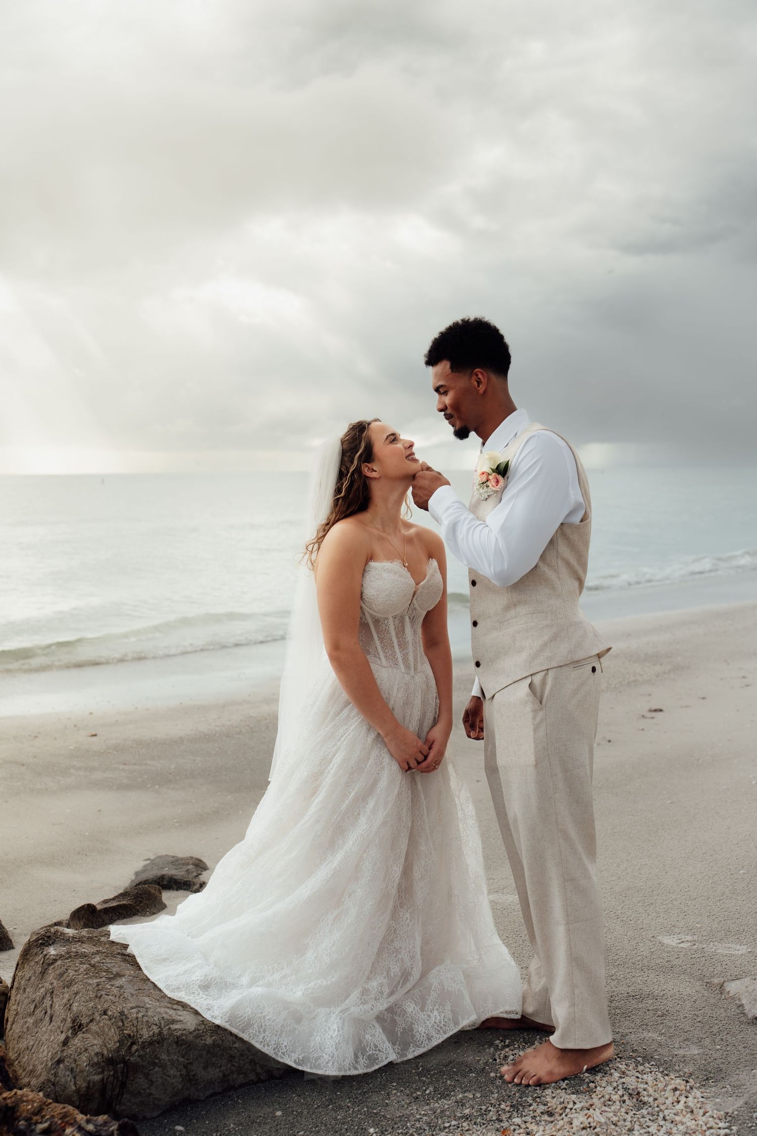 Bride and groom share a romantic moment on a beach, with ocean waves and cloudy skies in the background.