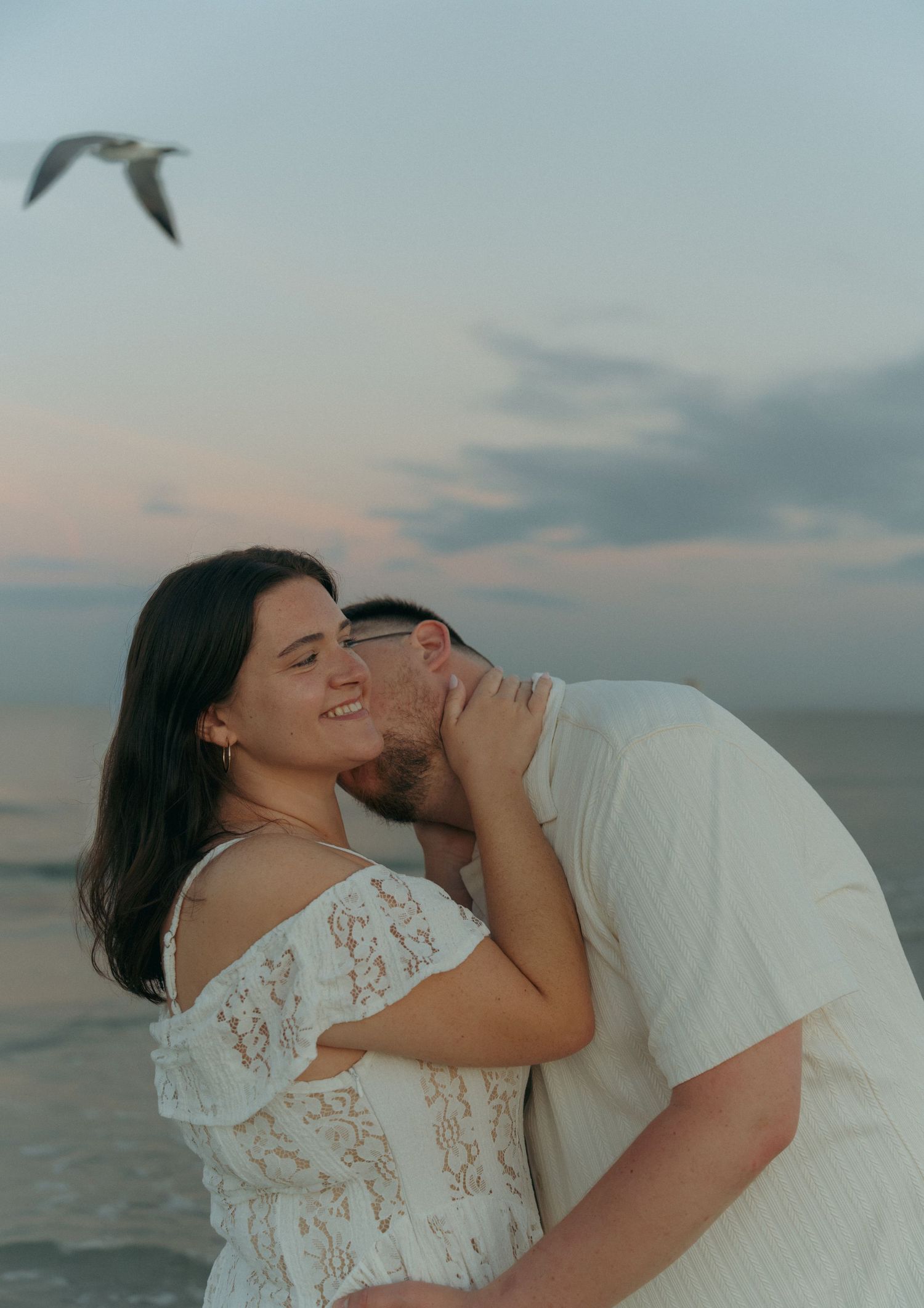Couple sharing a romantic beach moment at sunset, woman in white lace dress smiling as man kisses her neck.