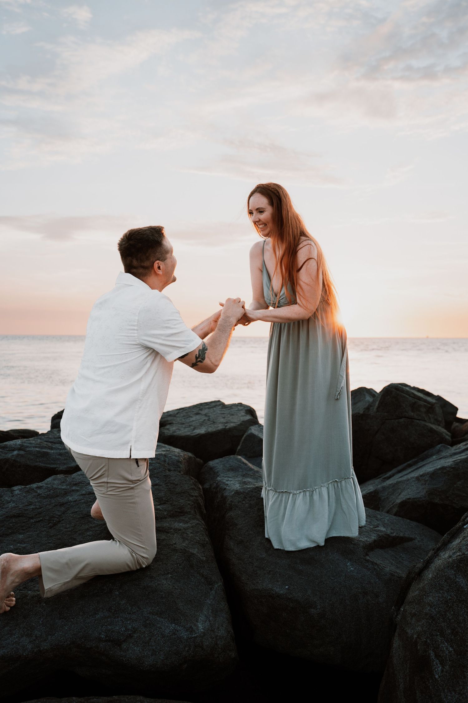 Man proposes to woman on rocky beach at sunset, both smiling as he kneels holding her hands.