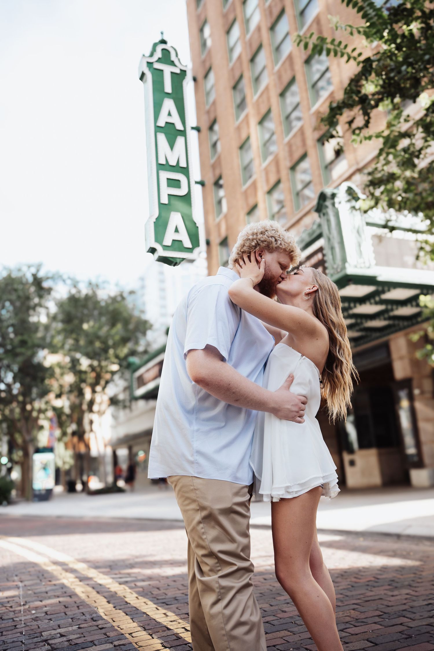 Couple embracing and kissing in front of the iconic Tampa Theatre marquee sign on a sunny day in Tampa, Florida.